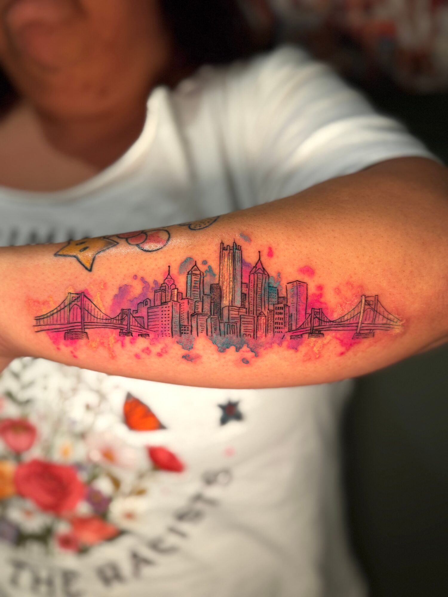 Tattoo of a city skyline with bridges and buildings on a person's arm, with a watercolor background.
