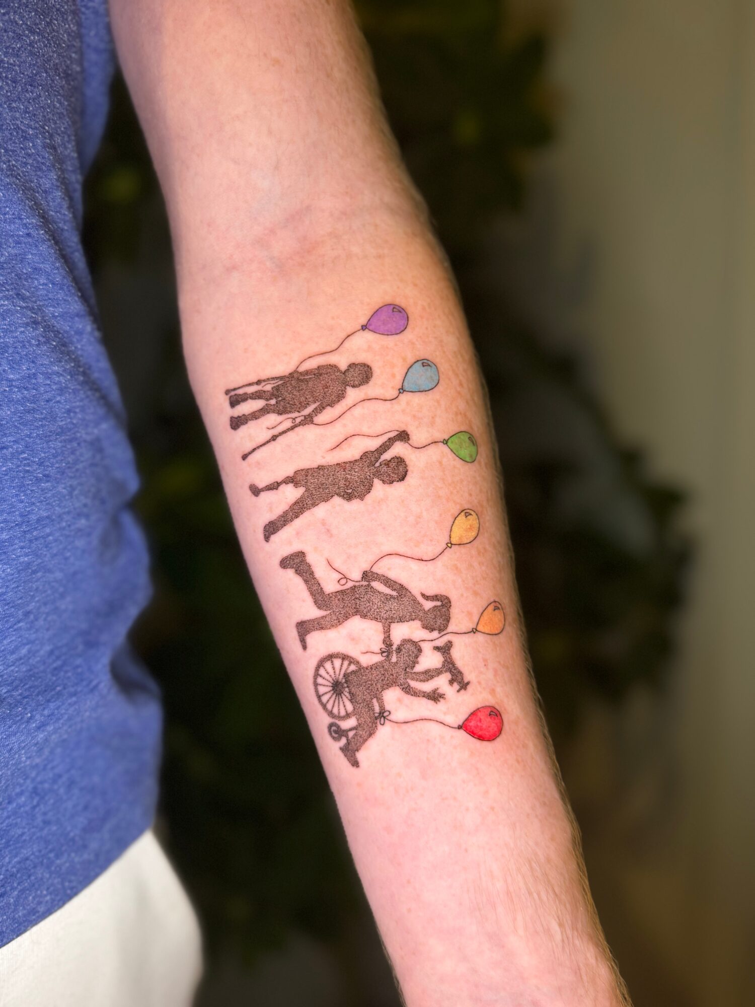 Forearm tattoo of five silhouettes of children holding balloons in different colors, with a bicycle at the bottom.