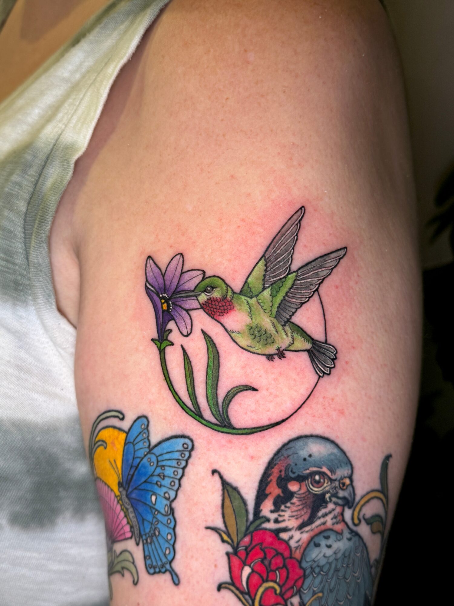 Hummingbird with purple flower, blue butterfly, and bird with red flower on arm, colorful tattoo, skin visible.