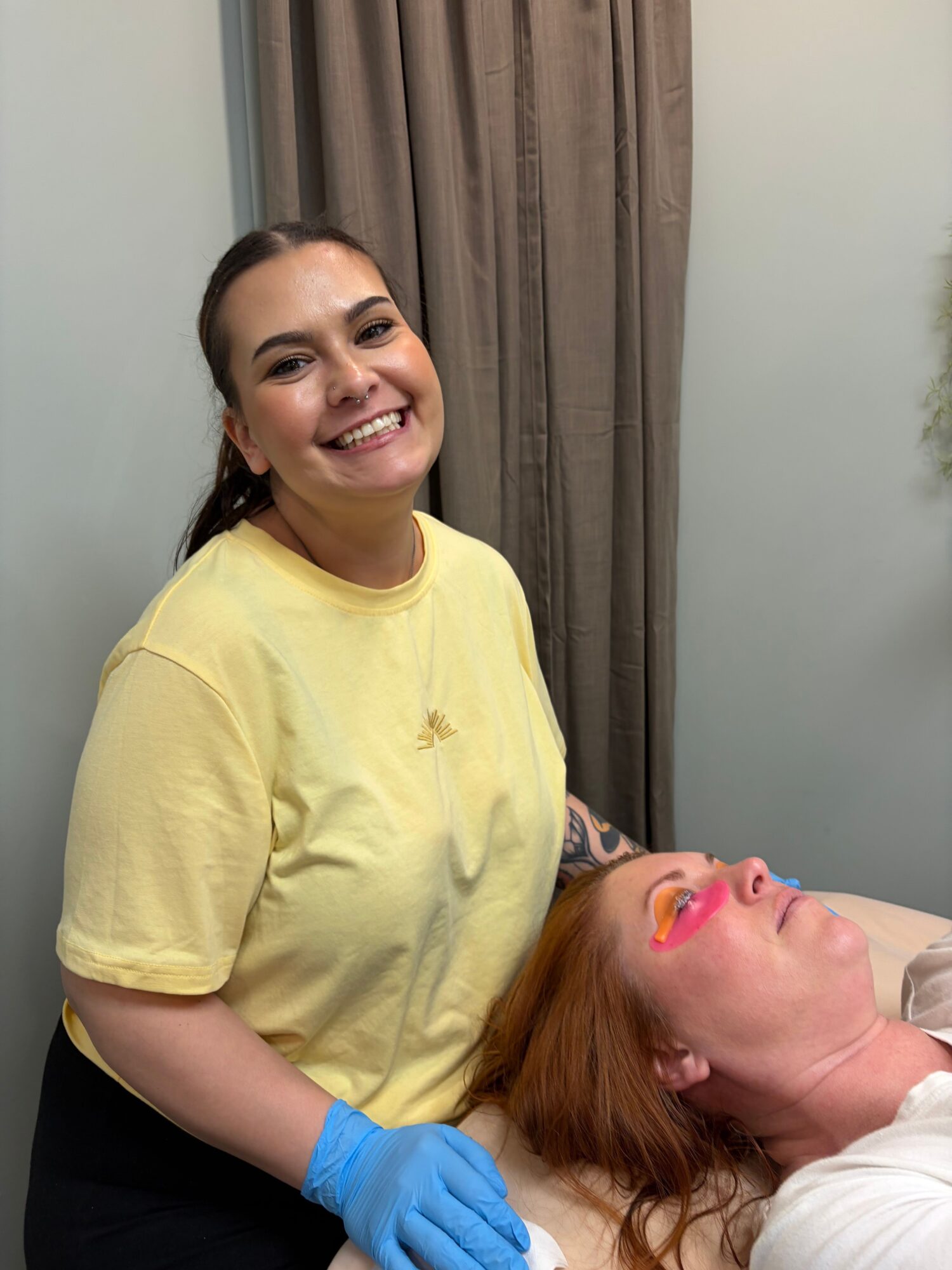 Two women smiling, one wearing a yellow shirt and blue gloves, the other lying down with a white shirt, in a room.