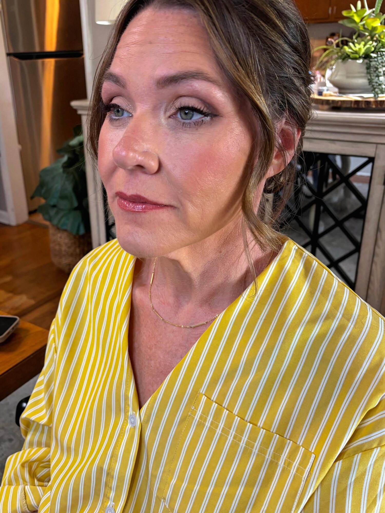 Woman with brown hair and blue eyes wearing a yellow and white striped shirt indoors, near a table with plants.