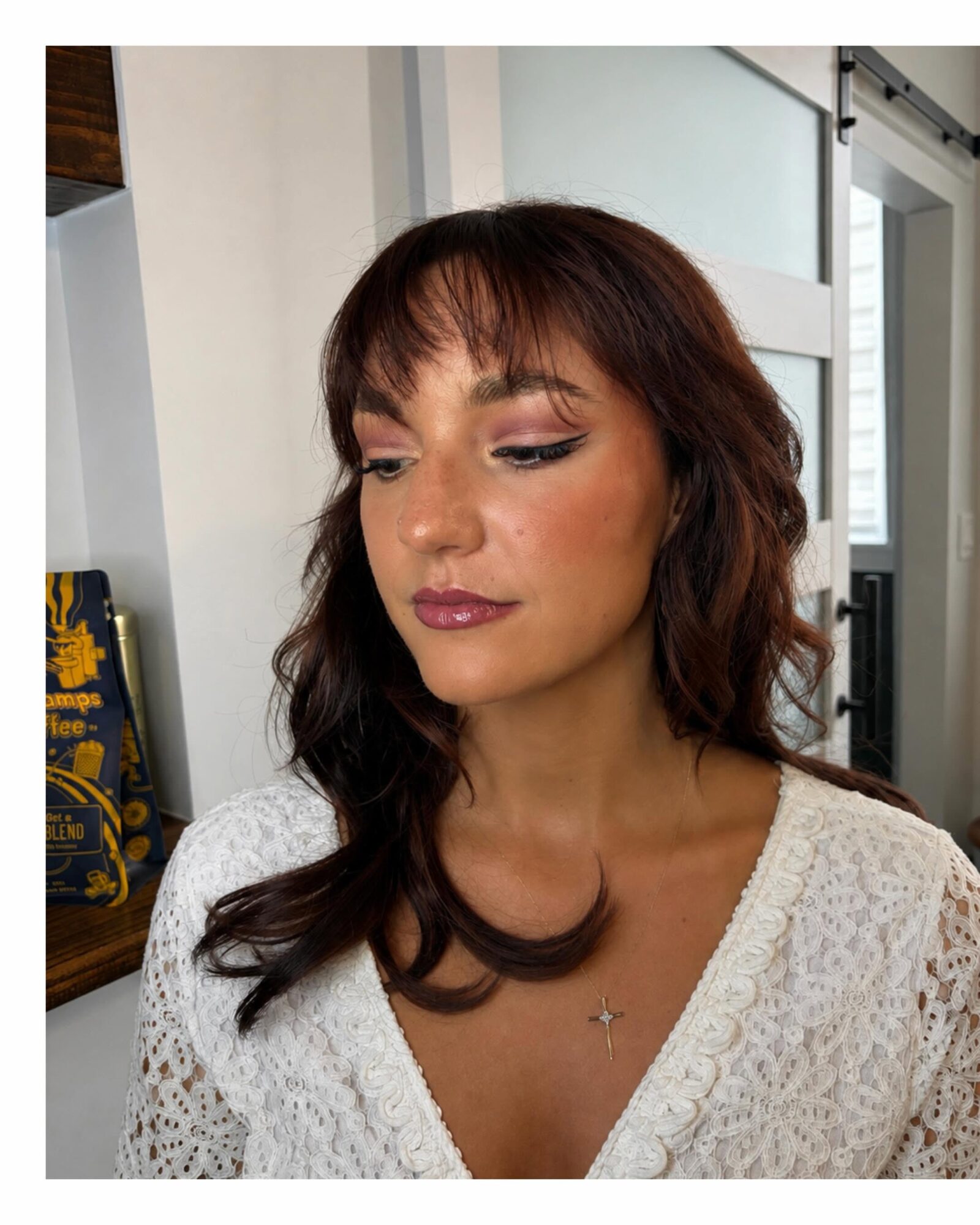 Woman with shoulder-length wavy brown hair and makeup, wearing a white textured top, looking down, indoors with a door and window behind.