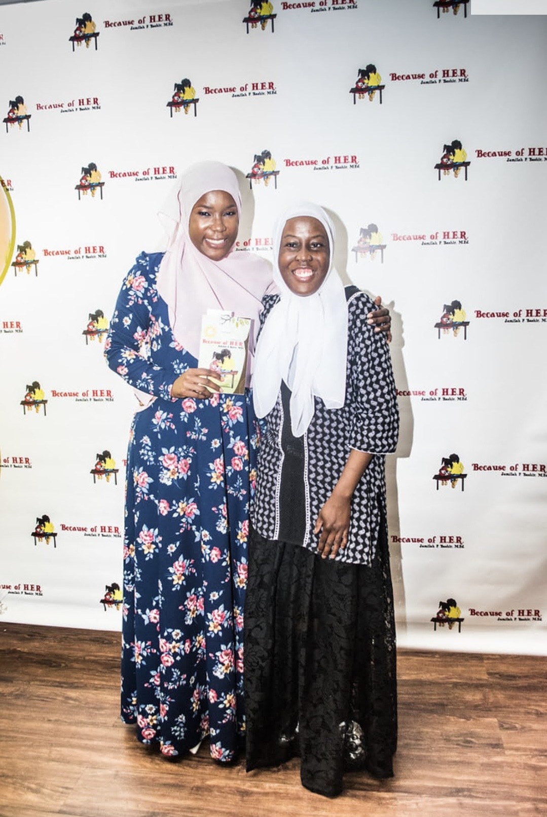 Two women in hijabs smiling and standing together in front of a backdrop with a horse logo and text, holding a book.