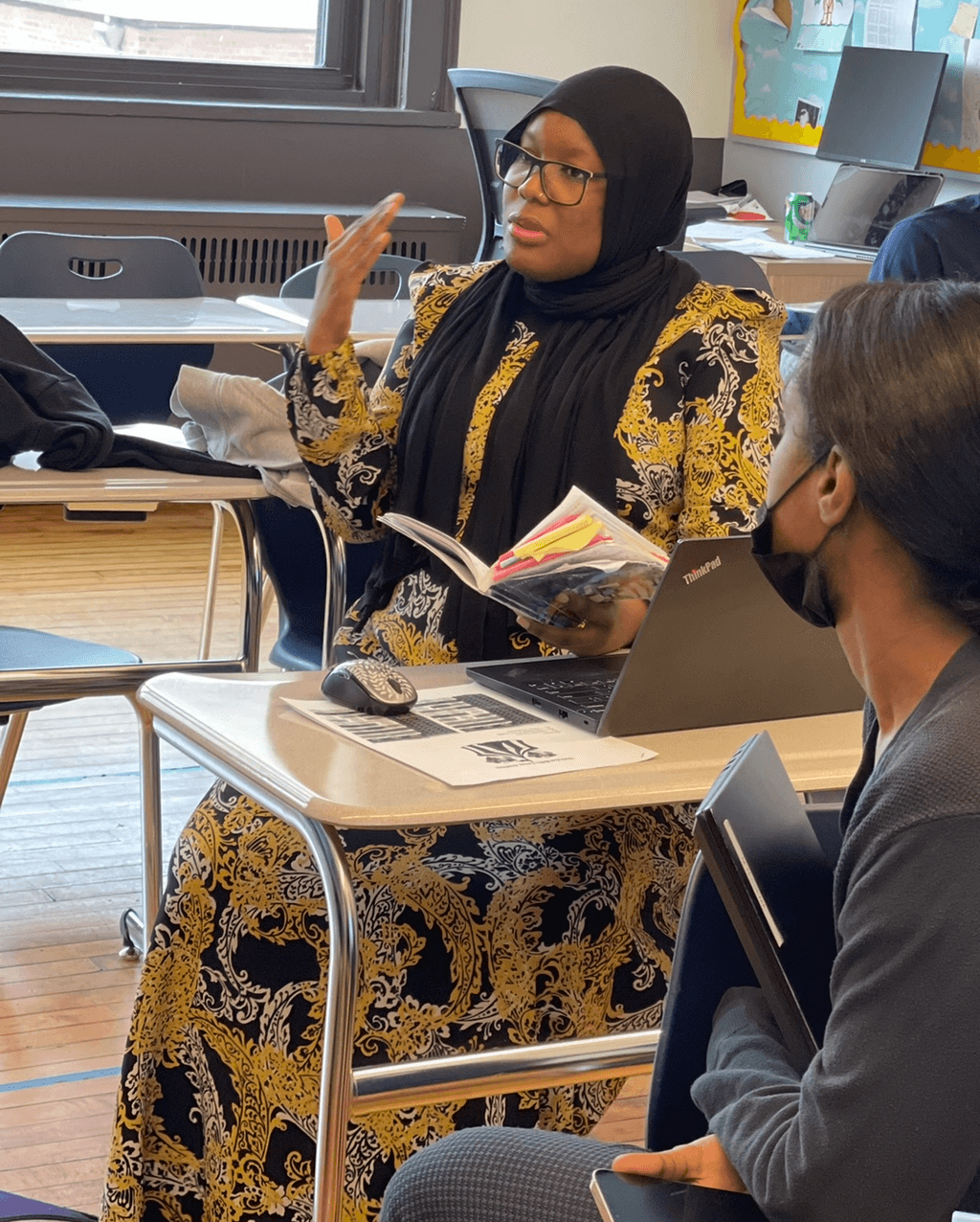 Woman wearing a black hijab and yellow patterned dress speaking in a classroom, holding a book, with students and desks around.