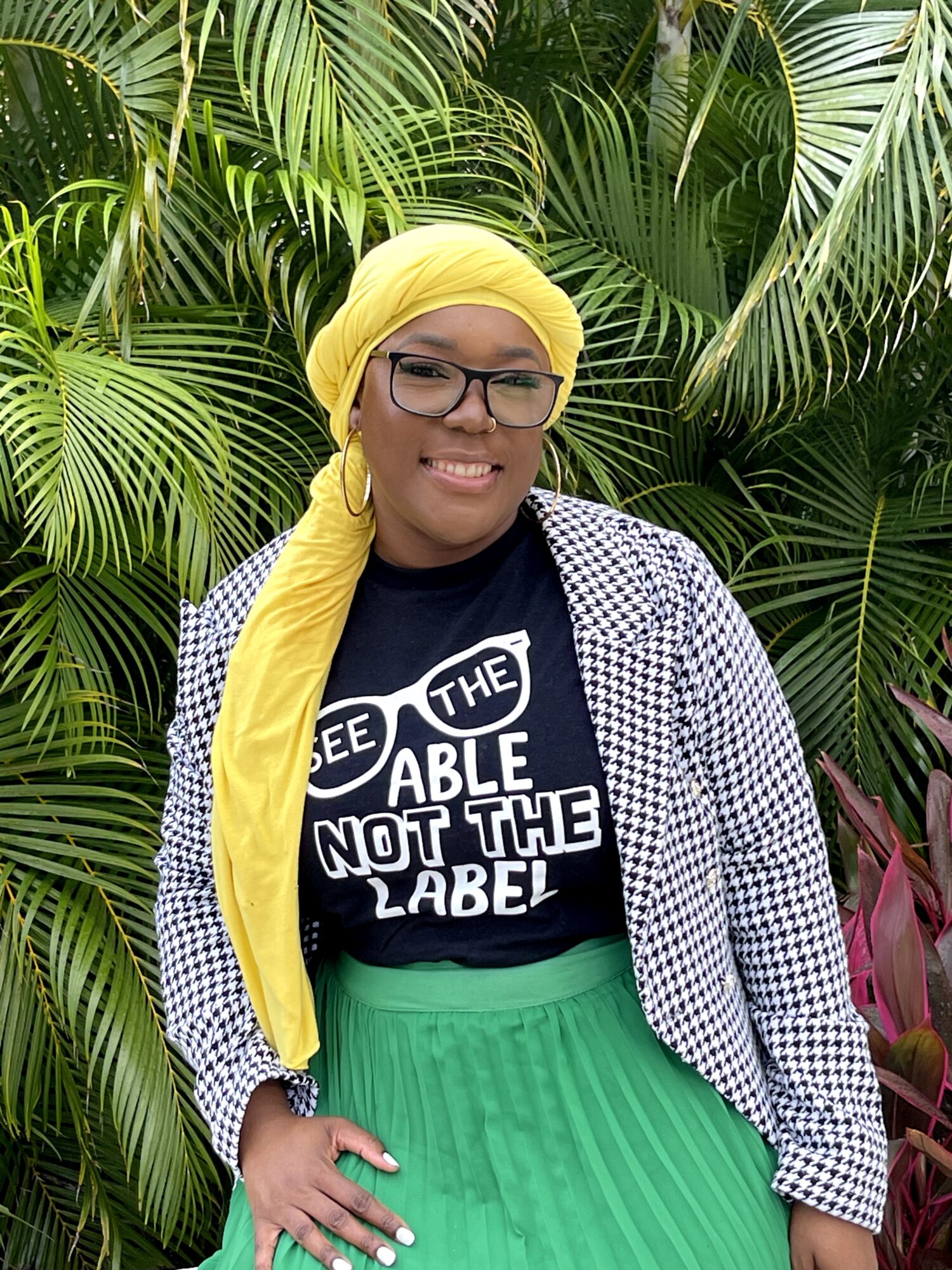 Woman wearing glasses, yellow headscarf, black T-shirt, and green skirt, standing in front of lush green plants.