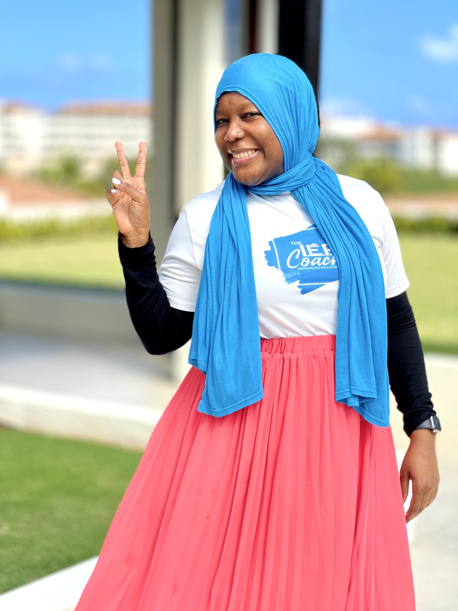 Smiling woman wearing a blue headscarf, white T-shirt, and pink skirt, making a peace sign with her right hand outdoors.