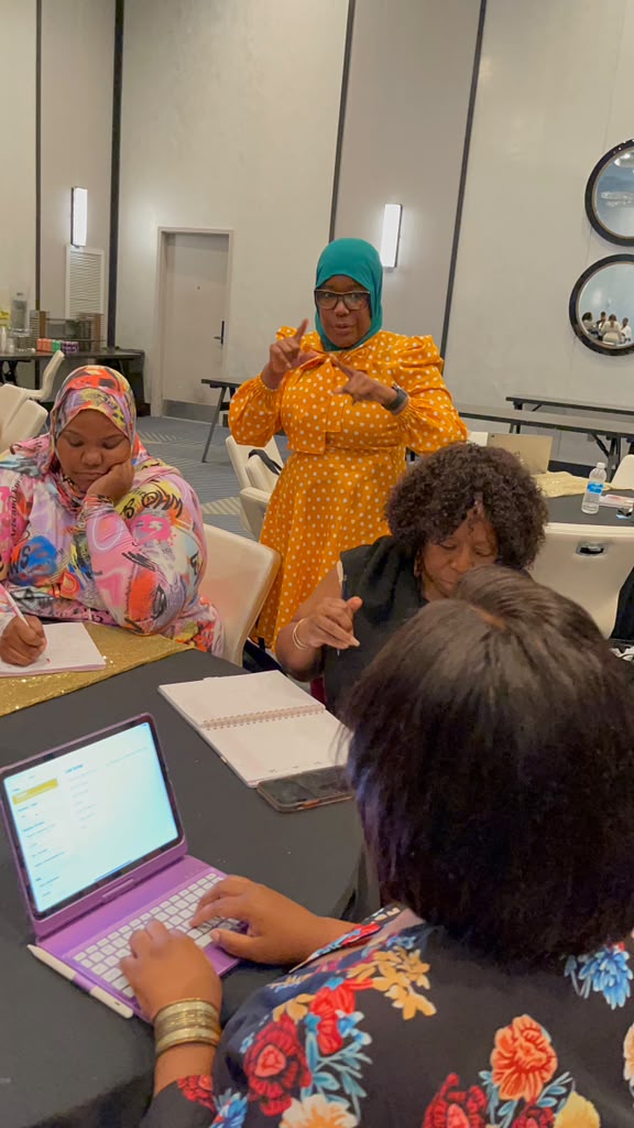 Three women sitting at a table with a laptop, notebooks, and a phone, in a modern room with mirrors and wall decor.