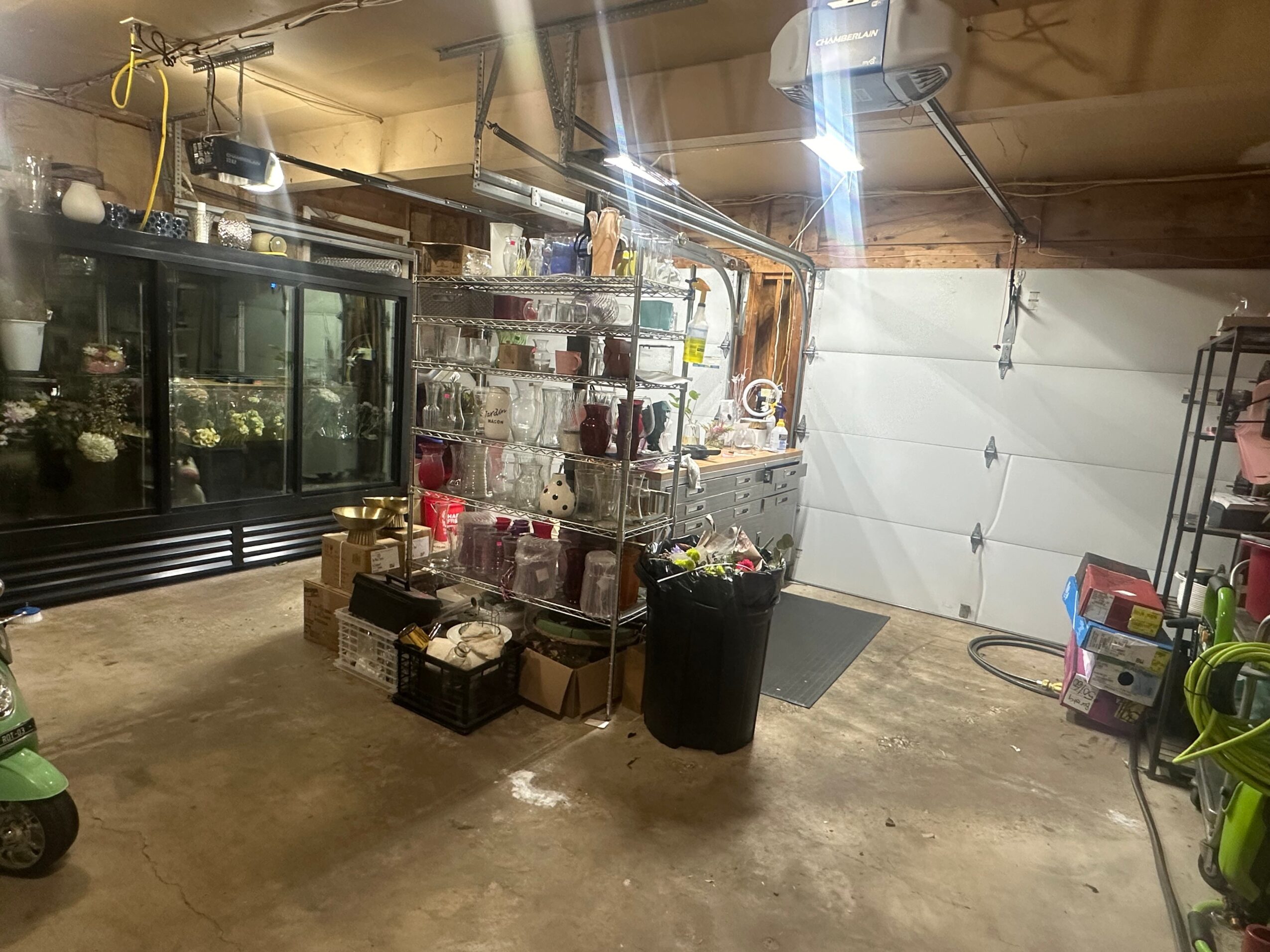 Garage with storage shelves, a large trash bin, and various items on the floor and shelves.