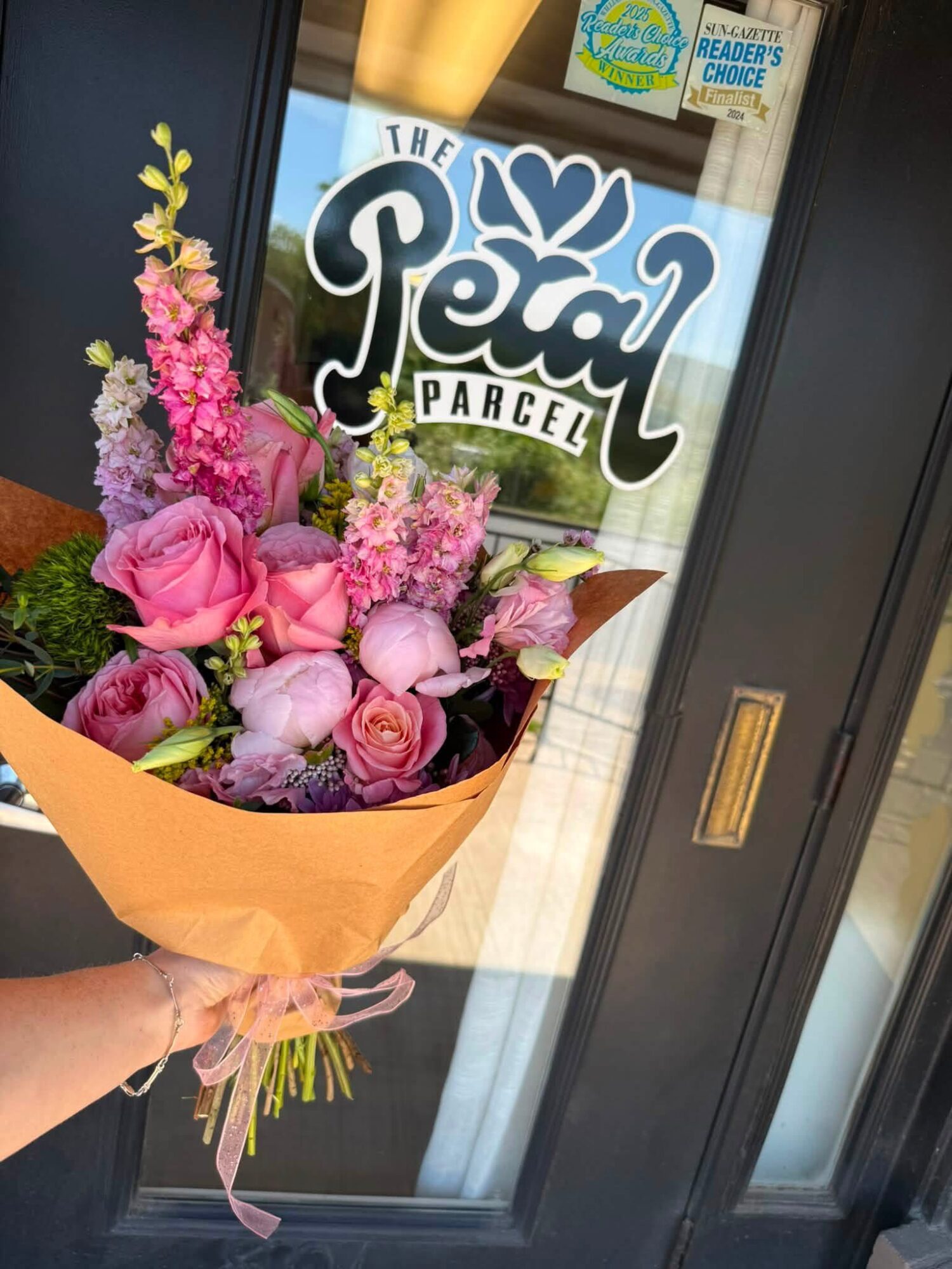 Person holding a bouquet of pink and purple flowers in front of a glass door with a sign reading 'The Petal Parcel'.
