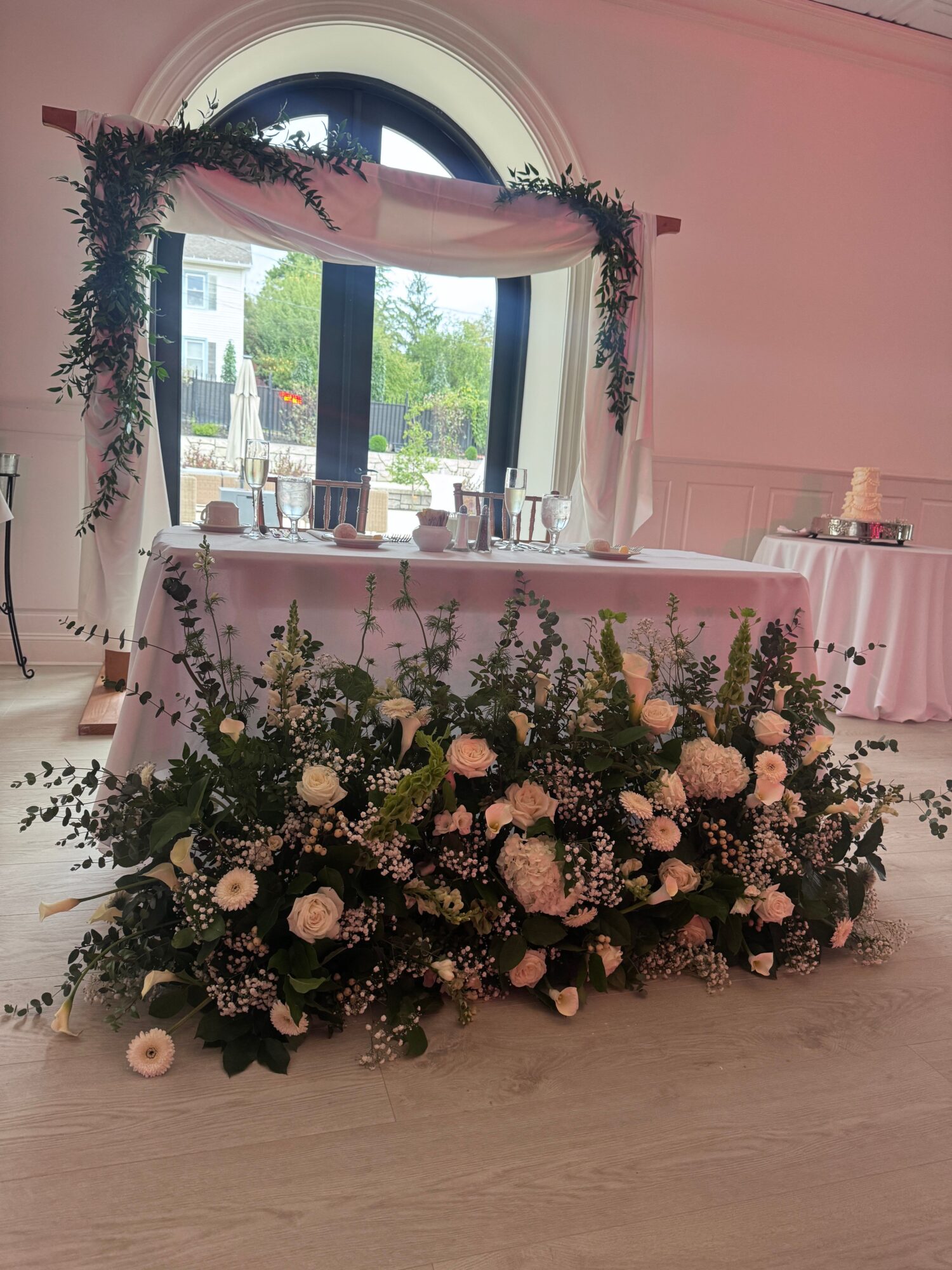 Wedding or event setup with a decorated arch, table, and floral arrangement near a window, pink walls, and wooden floor.
