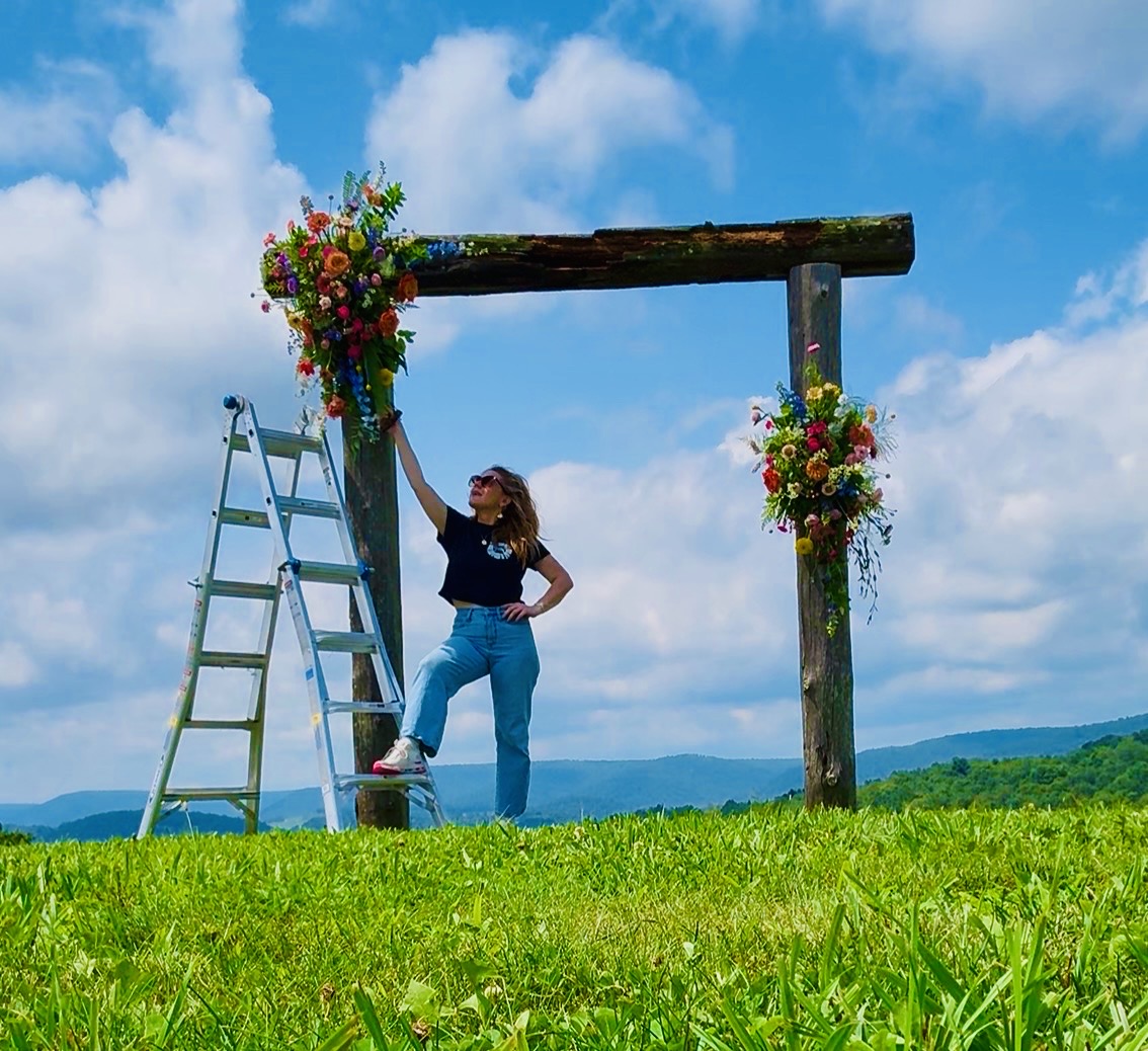 Woman standing next to a decorated wooden arch with flowers, on a grassy field under a blue sky with clouds.
