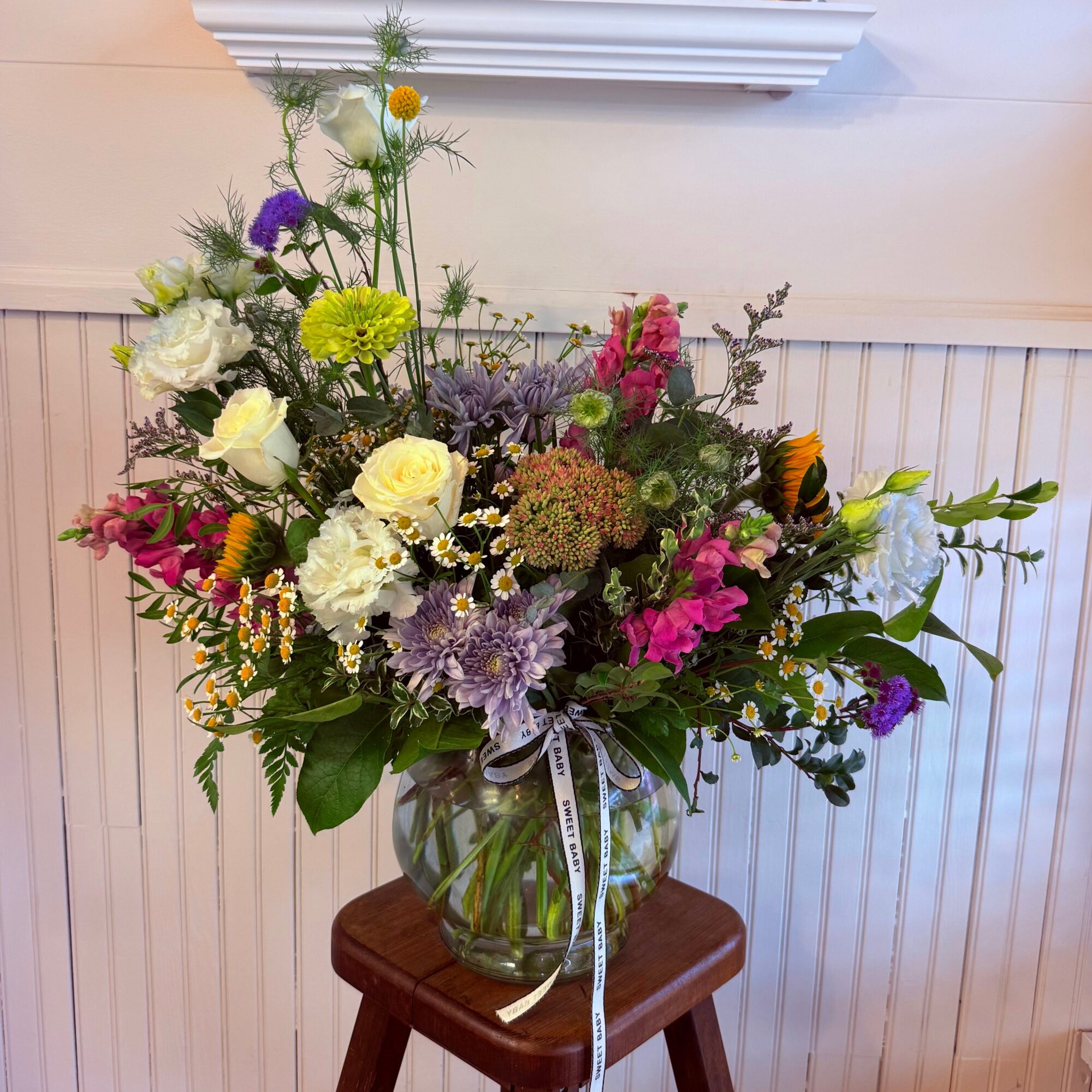 A colorful flower bouquet in a glass vase on a wooden stool, with various flowers and greenery, against a white wall.
