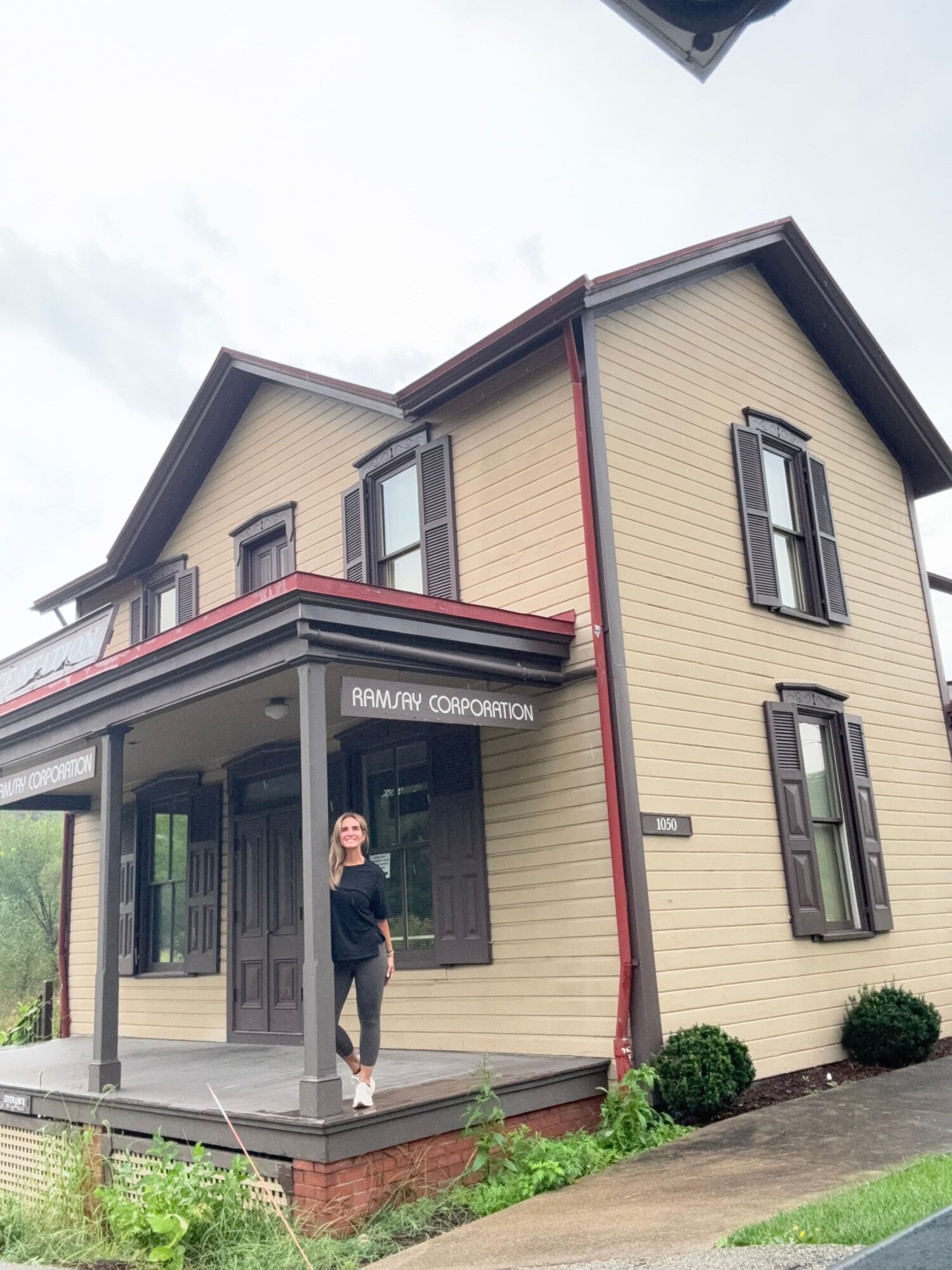 Two-story house with beige siding, black window frames, and a porch with a person standing outside, surrounded by grass and a garden bed.
