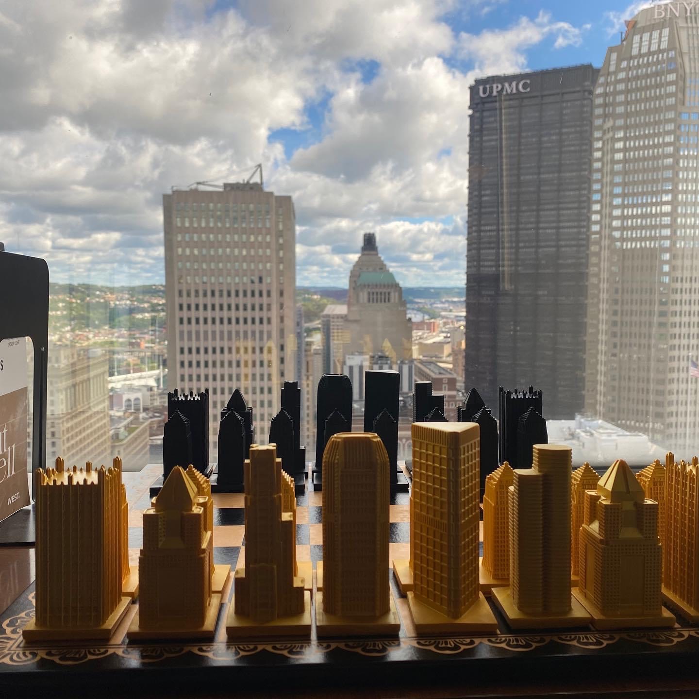 City skyline with tall buildings and a partly cloudy sky, viewed from a high window with model buildings in foreground.