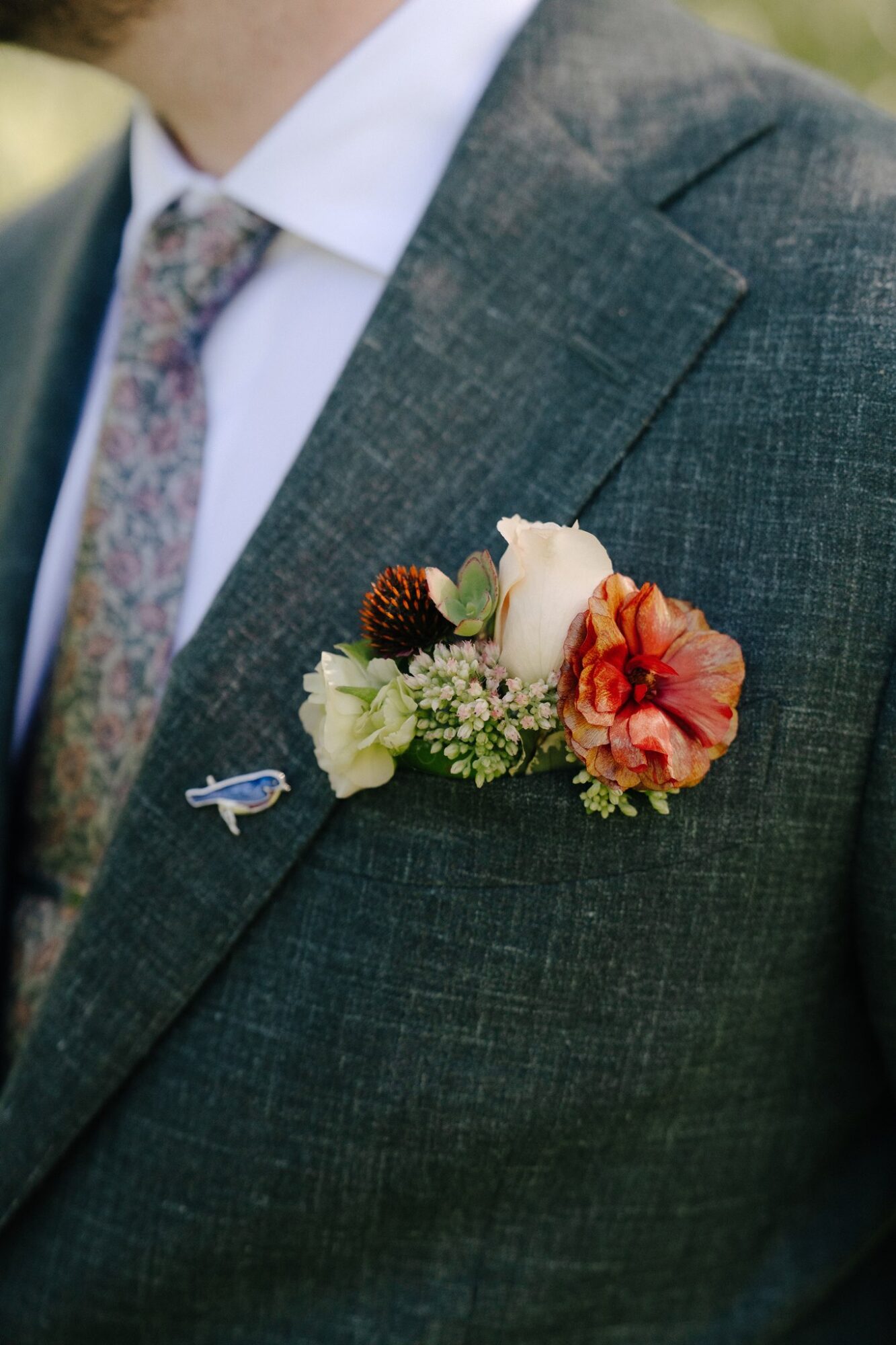 Close-up of a man's suit with a boutonniere featuring a white rose, orange flower, pine cone, and greenery on the lapel.