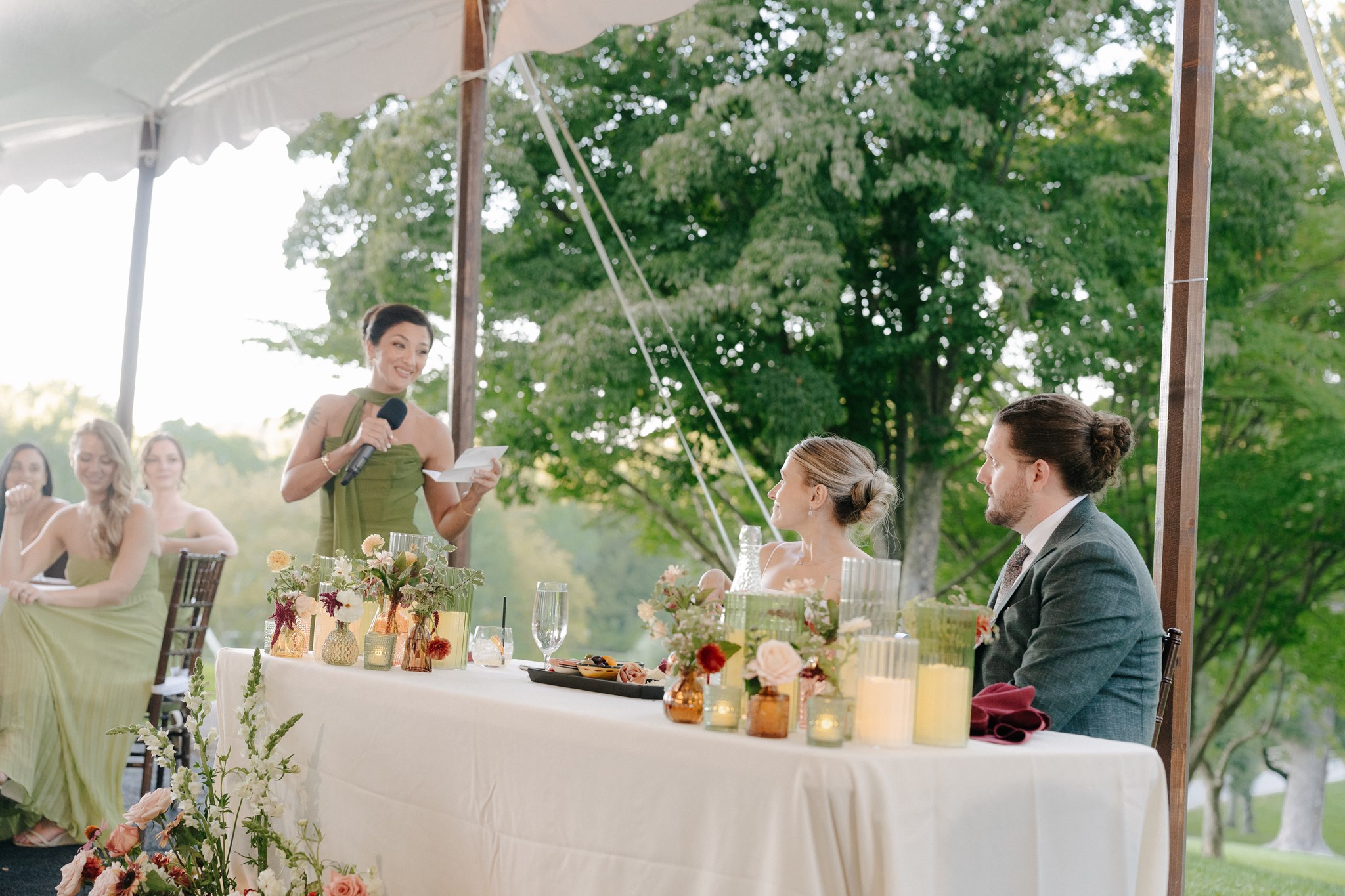 People sitting at a table outdoors under a tent, with trees in the background, celebrating with food and drinks.