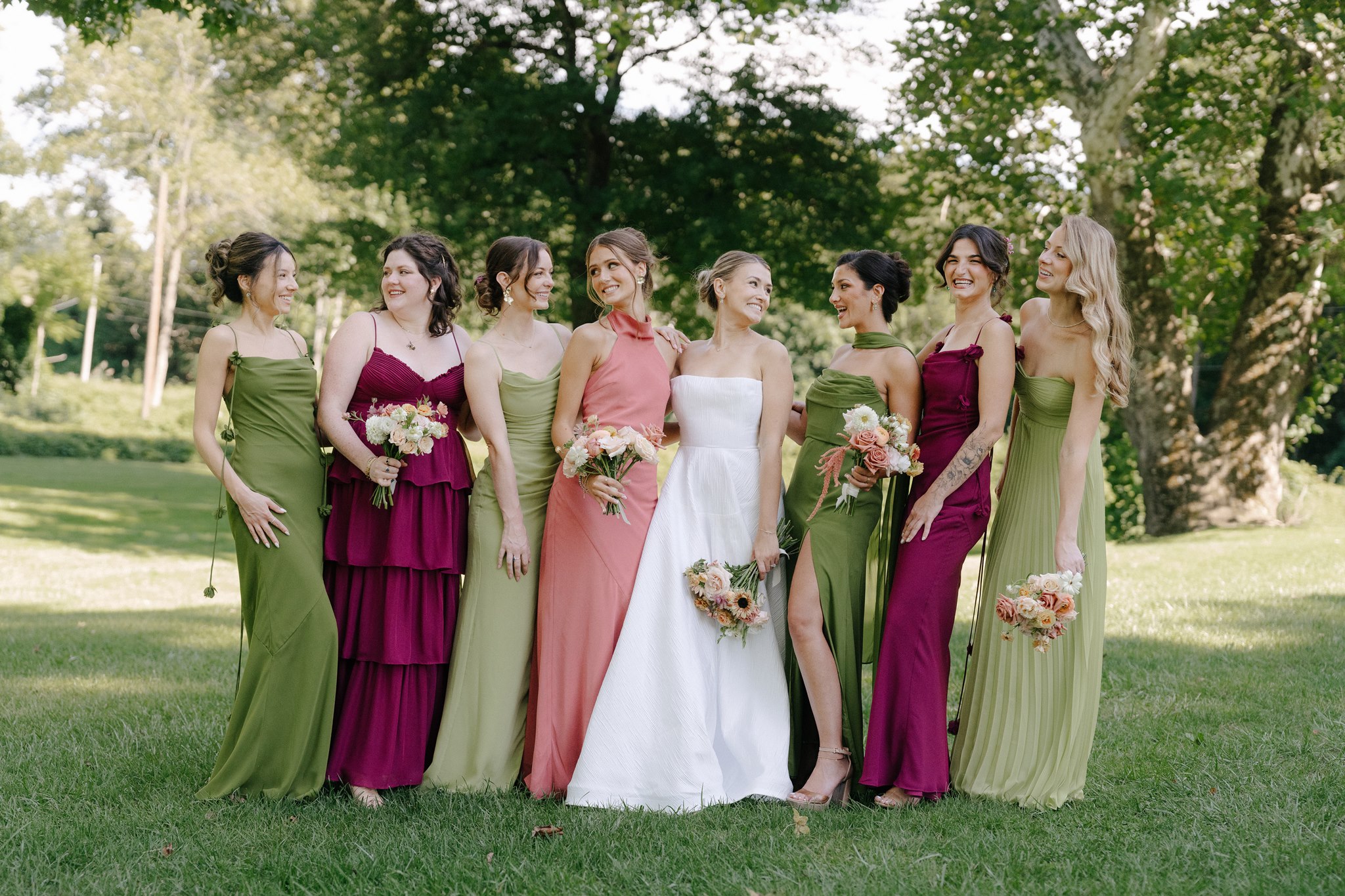 Group of seven women, including a bride in a white dress, standing outdoors on grass with trees in background.