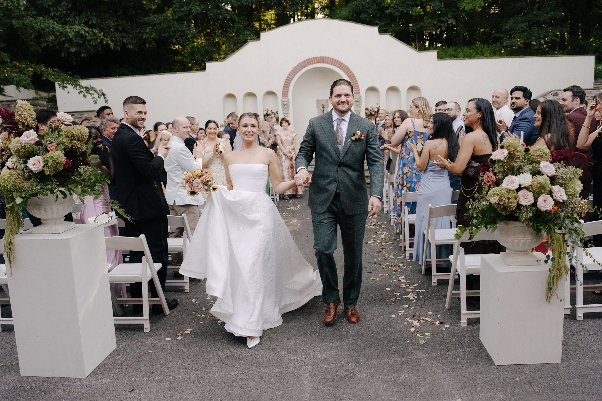 Bride and groom walking down aisle with guests on both sides, floral arrangements, outdoor wedding setting.