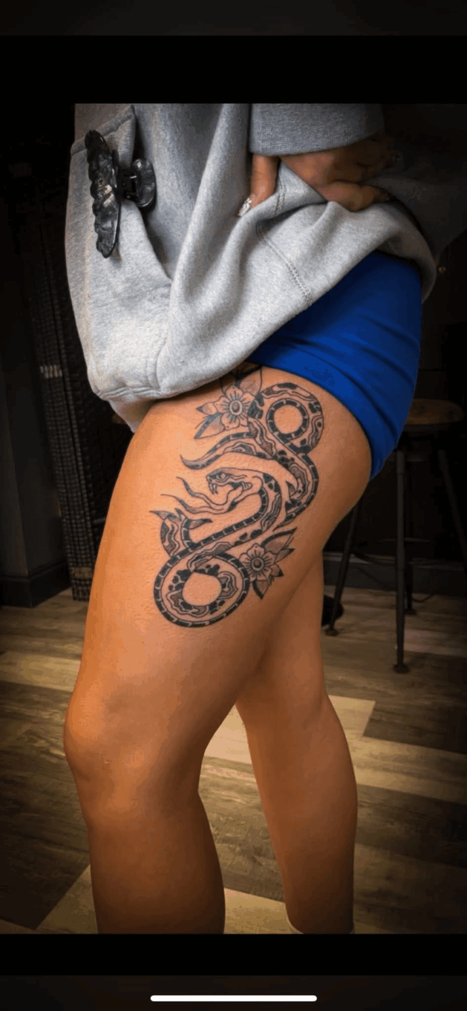 Person with a large snake tattoo on thigh, wearing a blue skirt and gray sweater, standing indoors.