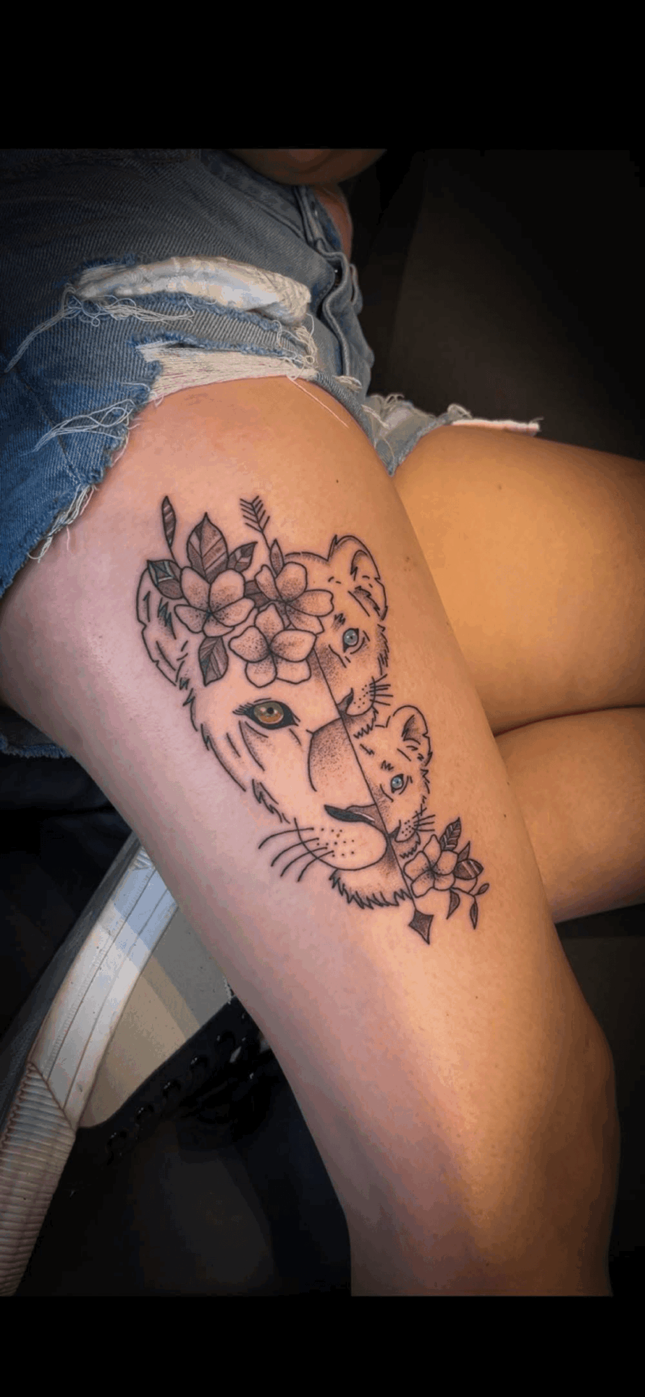 Tattoo of a tiger's face with floral elements on a person's thigh, wearing ripped denim shorts.