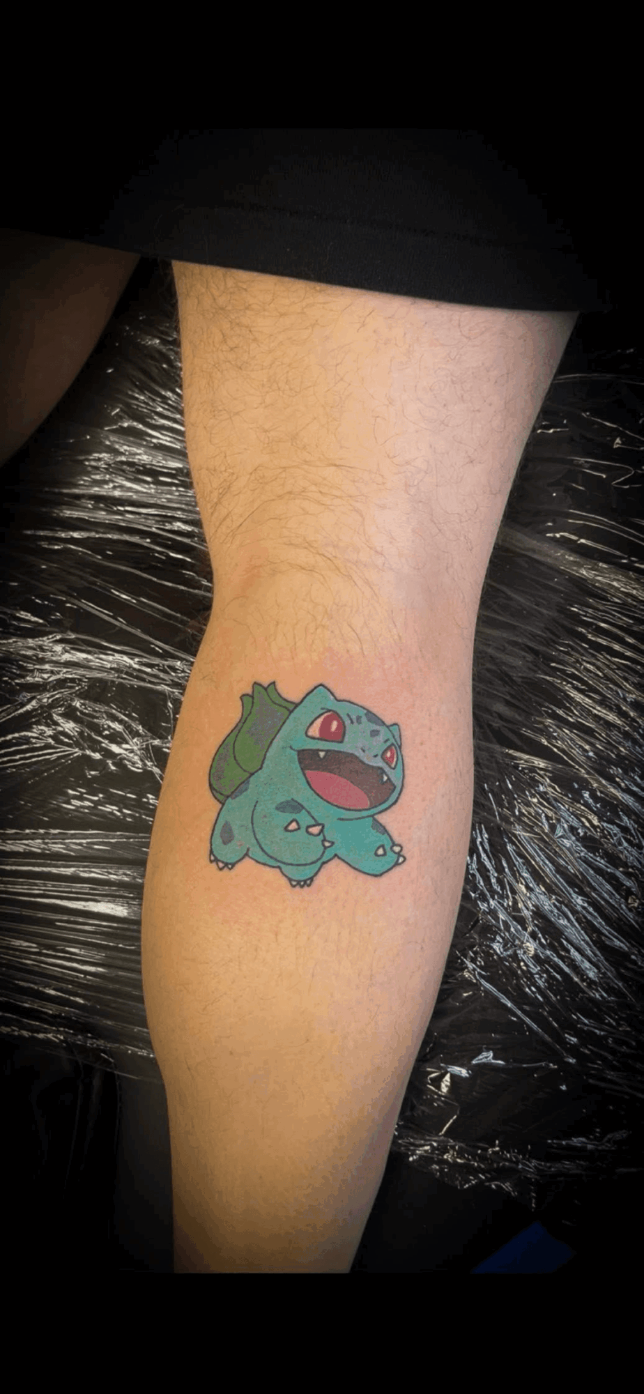 Colorful cartoon dinosaur tattoo on person's leg, with a green body, purple mouth, and small arms, on a light skin tone.