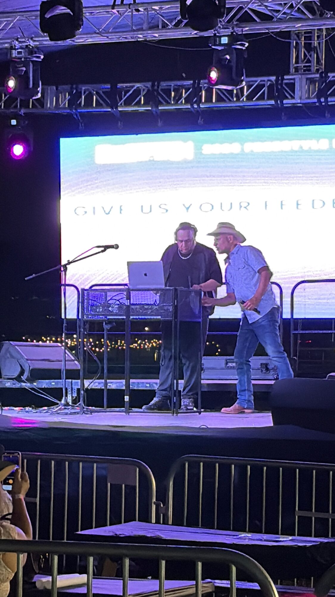 Two people on stage, one handing a document to the other, with a large screen and stage lighting behind them.