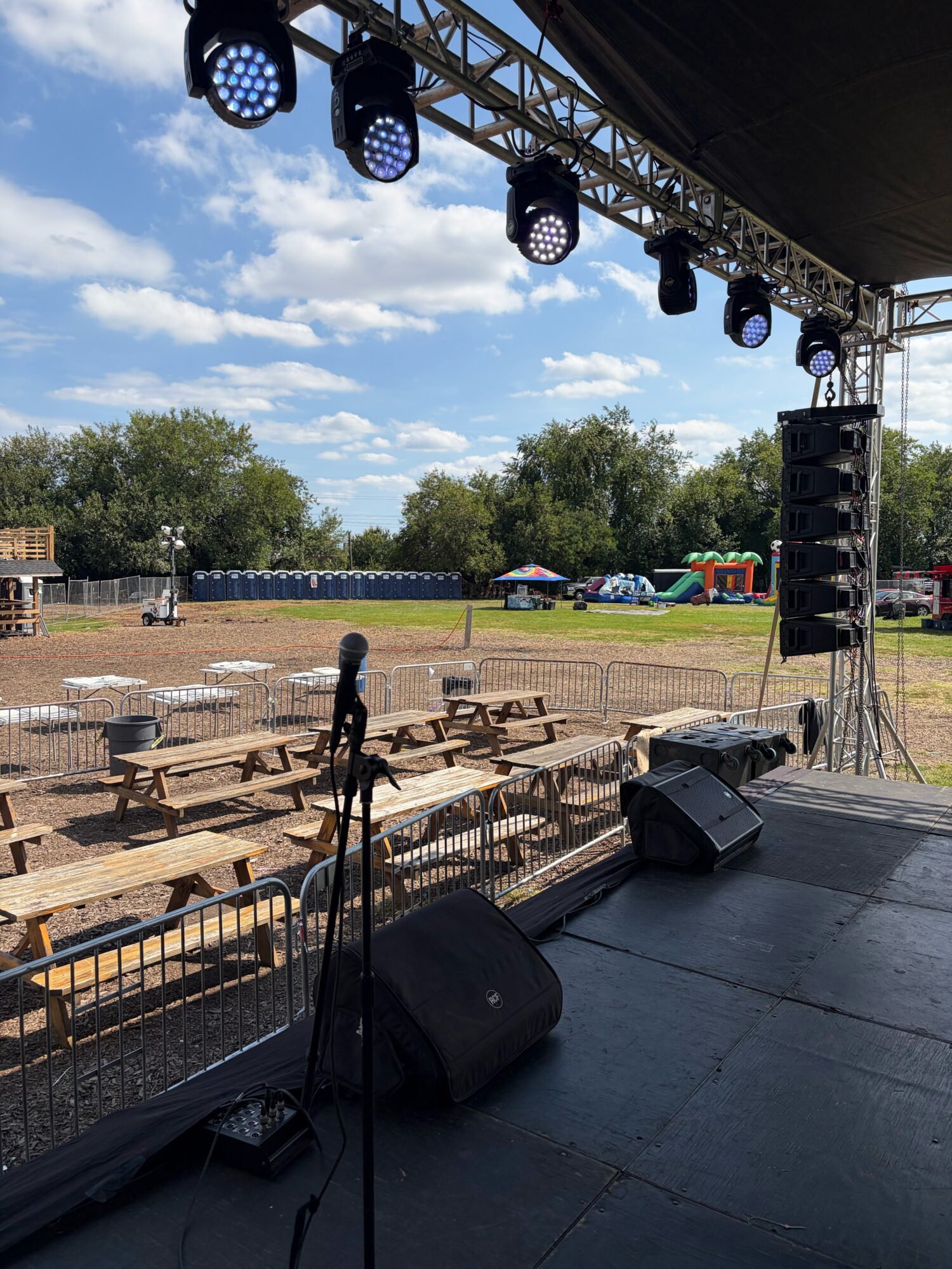 Stage with speakers and lighting, outdoor event setup with empty benches and a field, blue sky with clouds.