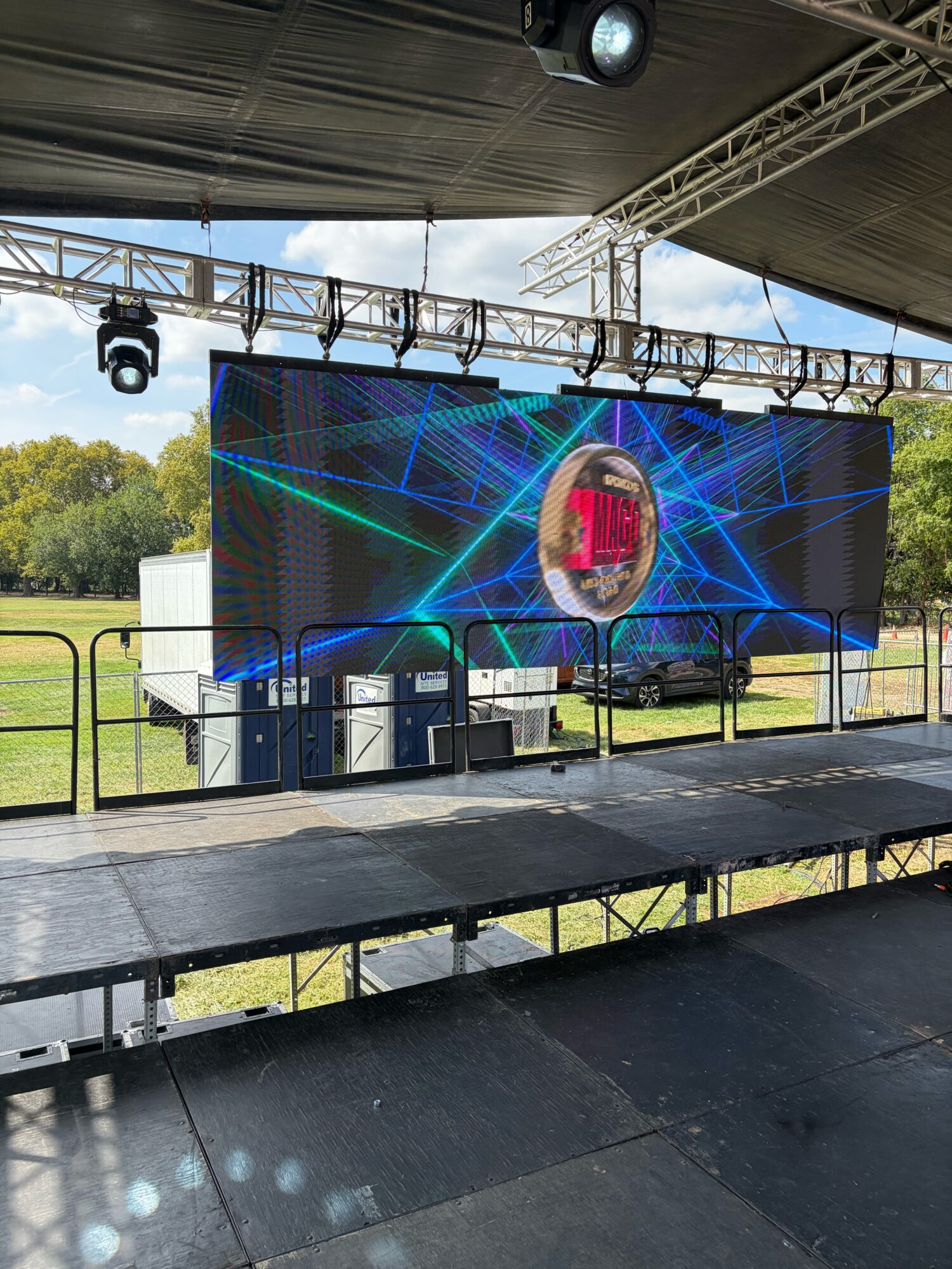 Empty outdoor stage with large LED screen, speakers, and lighting equipment, set up in a grassy field with trees in background.