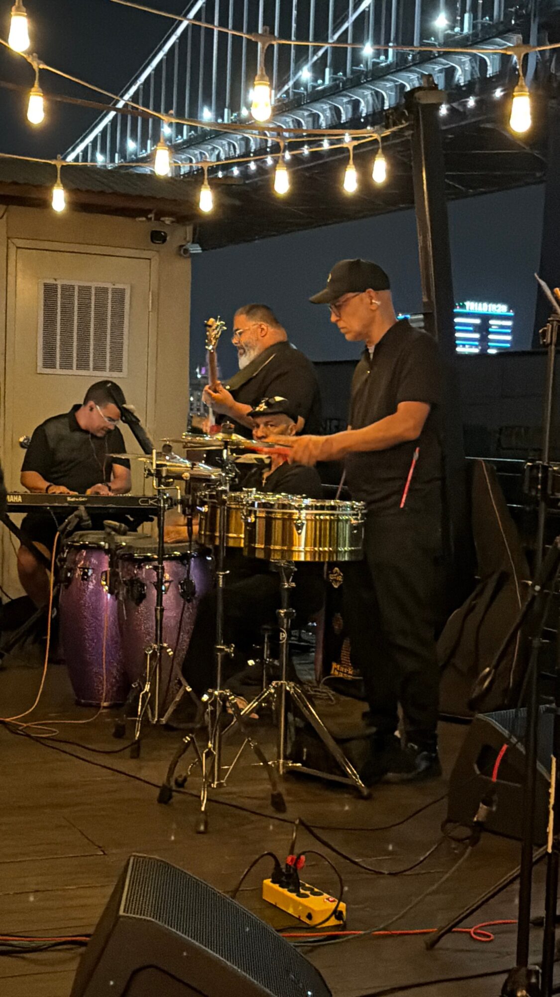 Three musicians perform on stage with drums, congas, and percussion instruments under string lights at night.