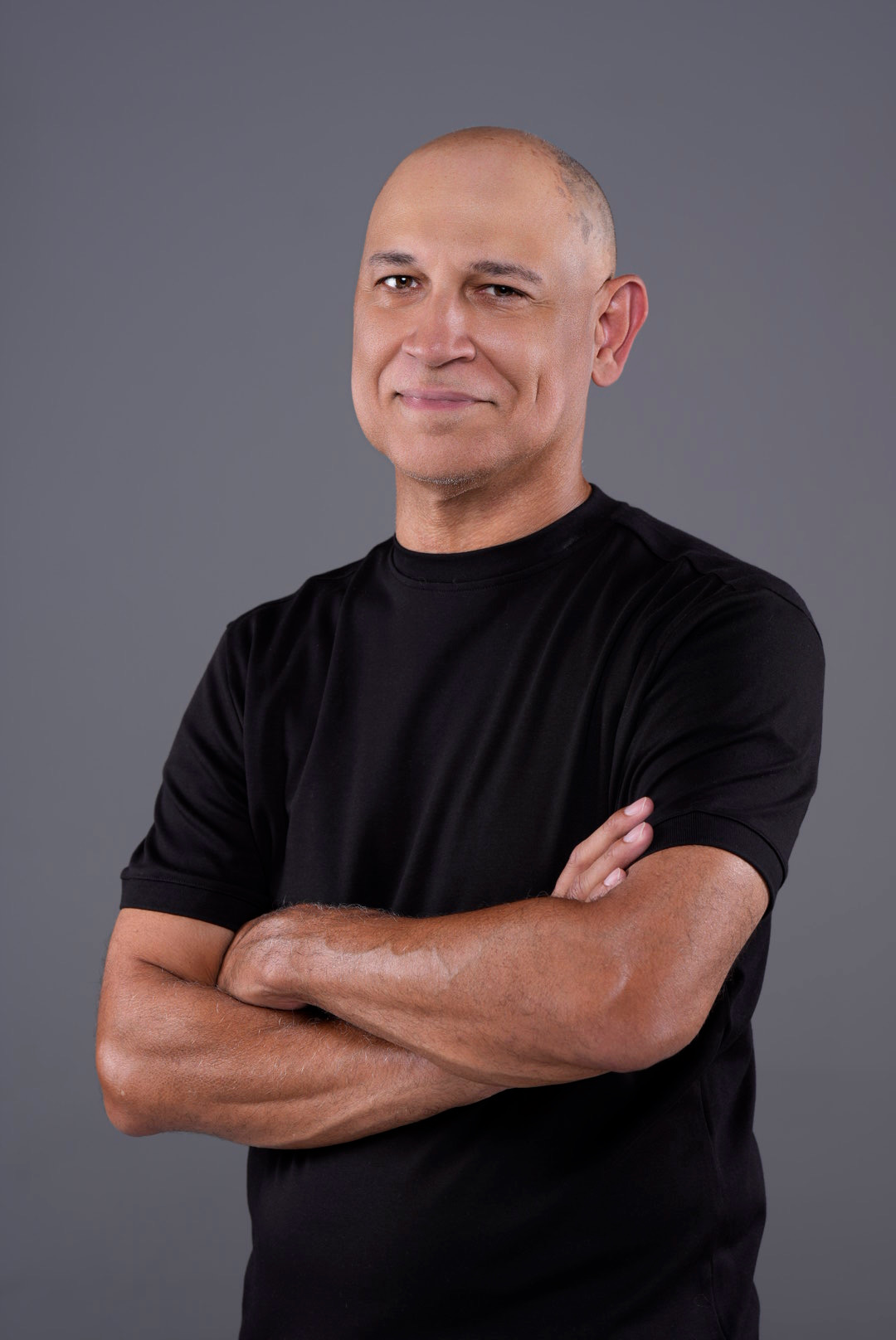 Man with crossed arms wearing a black shirt, smiling slightly, against a gray background.