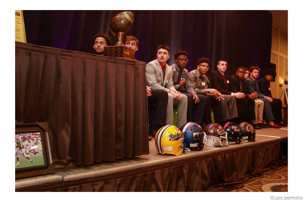 Group of people sitting on a stage with football helmets in front, a trophy, and a small framed picture, with a dark curtain background.
