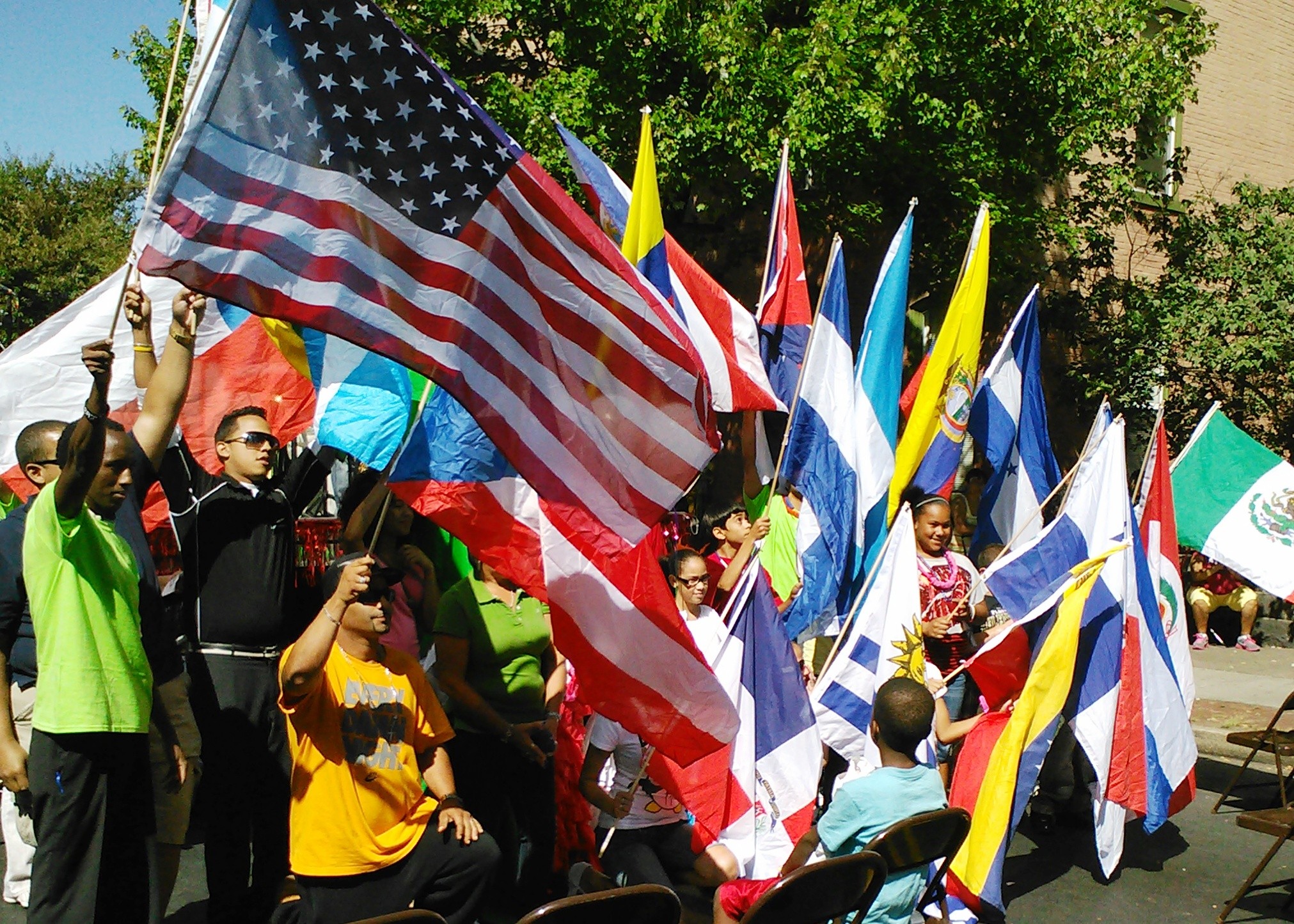 Group of people holding various national flags outdoors, with trees and buildings in the background.