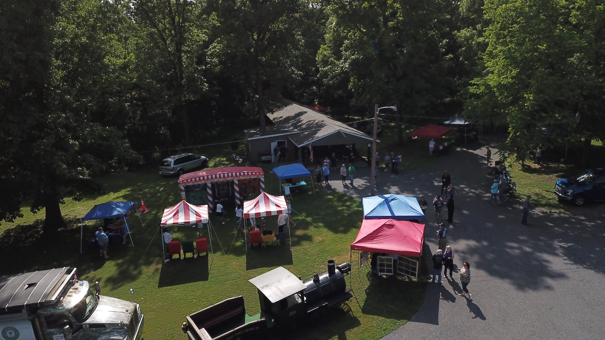 Outdoor event with tents, people, and vehicles in a green park area, trees in the background.