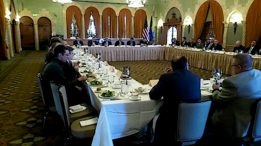 People seated around a large U-shaped table in a decorated room with arched windows and curtains.