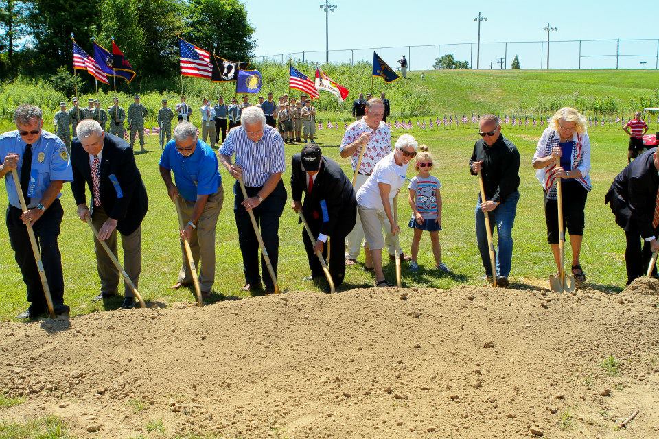 Group of people planting a tree outdoors with American flags in the background, on a sunny day.