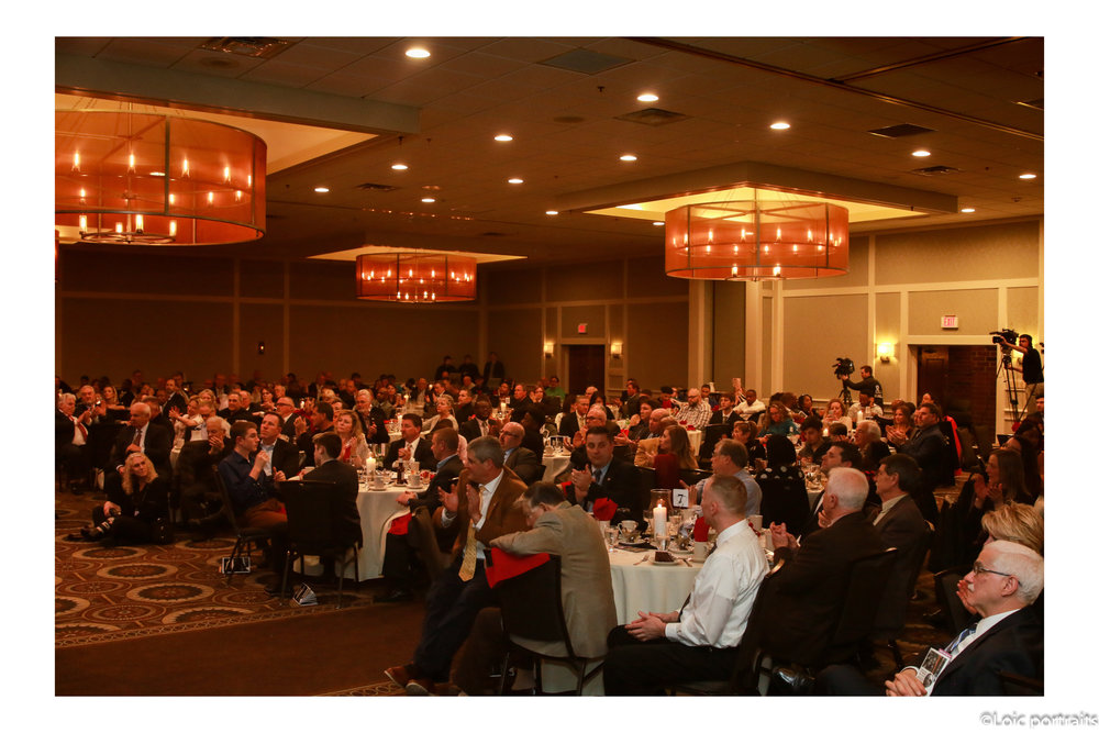 Large banquet hall filled with seated guests at round tables, warm lighting, and chandeliers, during an event or conference.