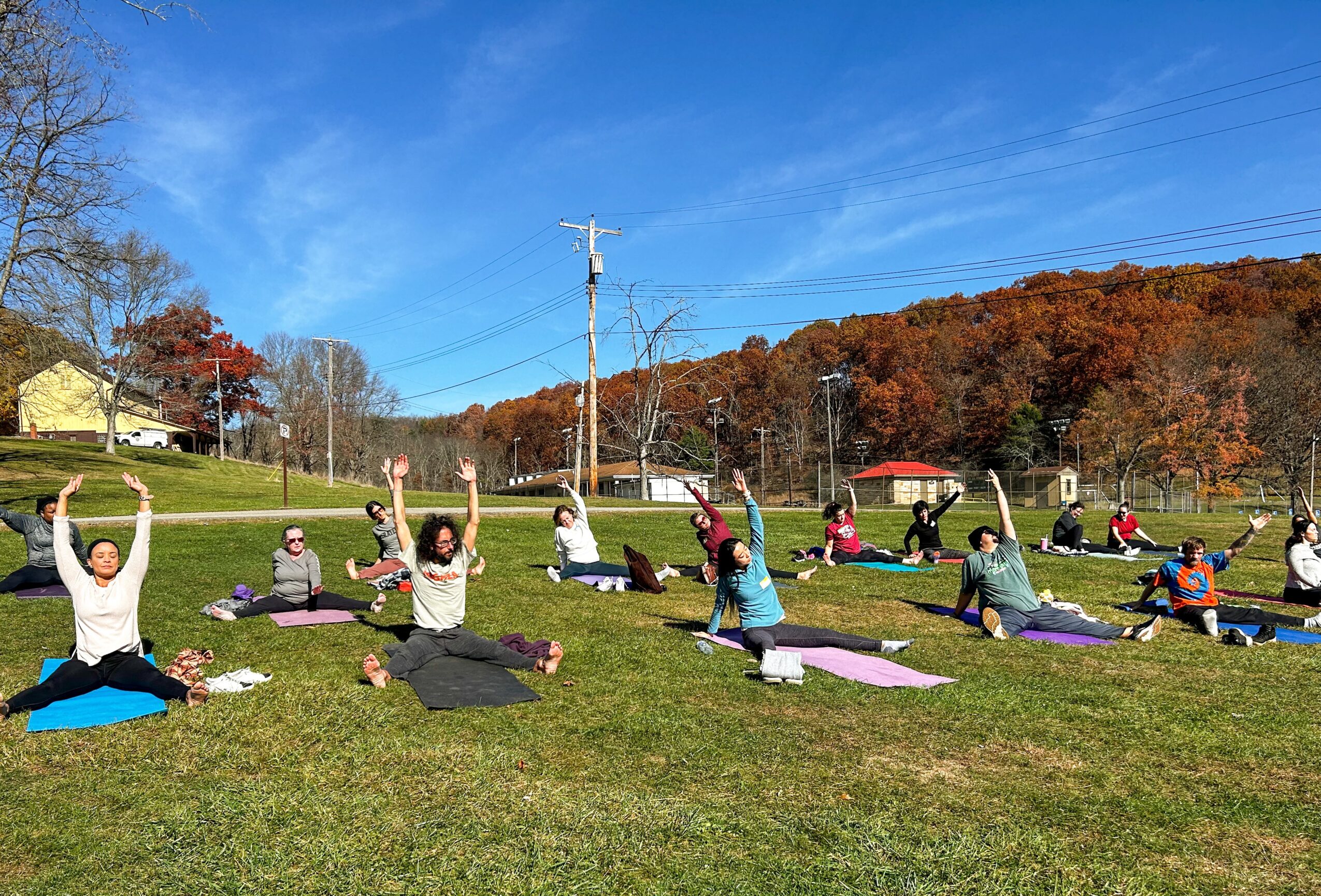 Group of people practicing yoga outdoors on a grassy field with trees and hills in the background, under a blue sky.