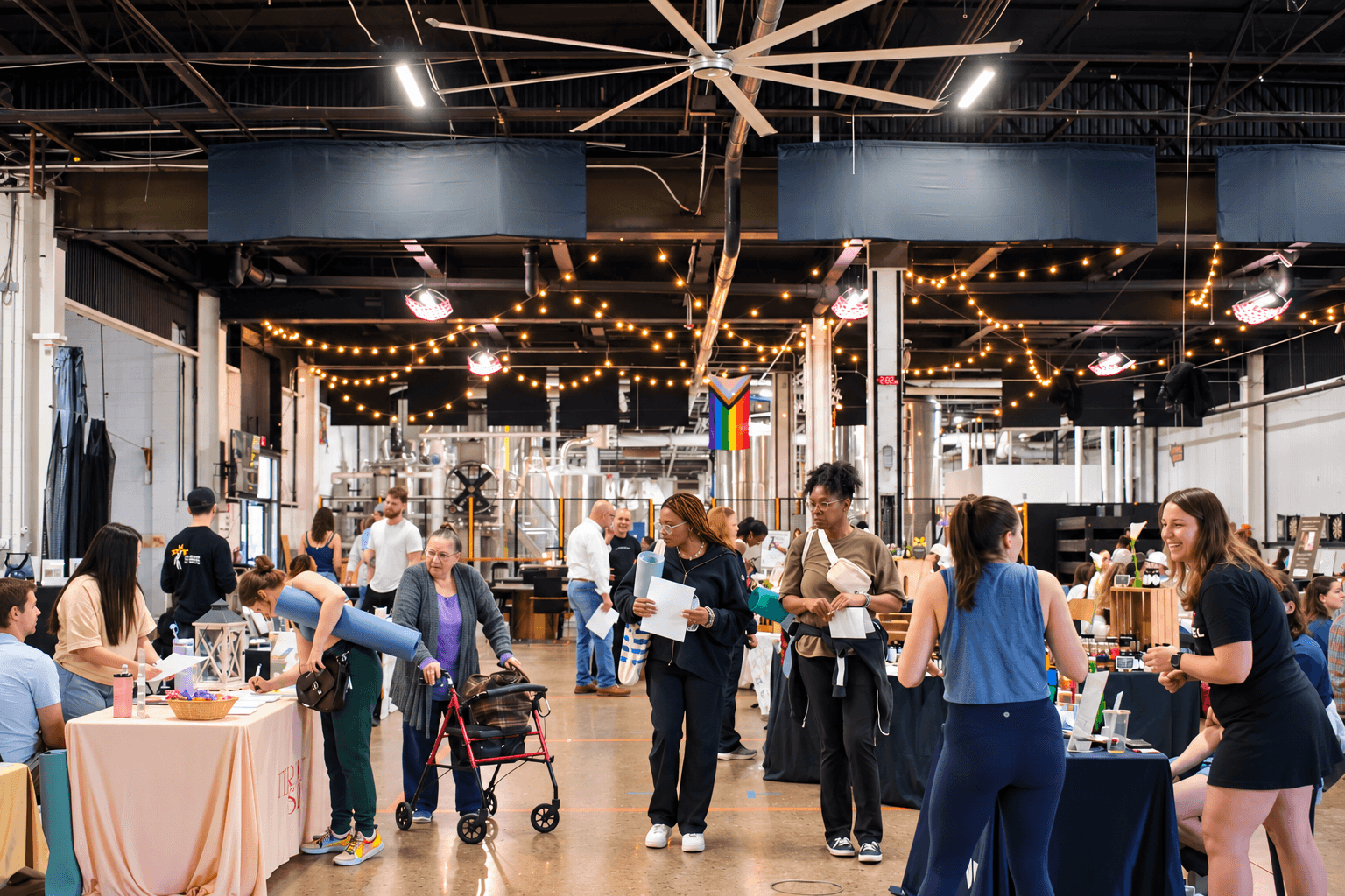 People gather in a spacious indoor venue with string lights, tables, and a bar area, engaging in conversations and activities.