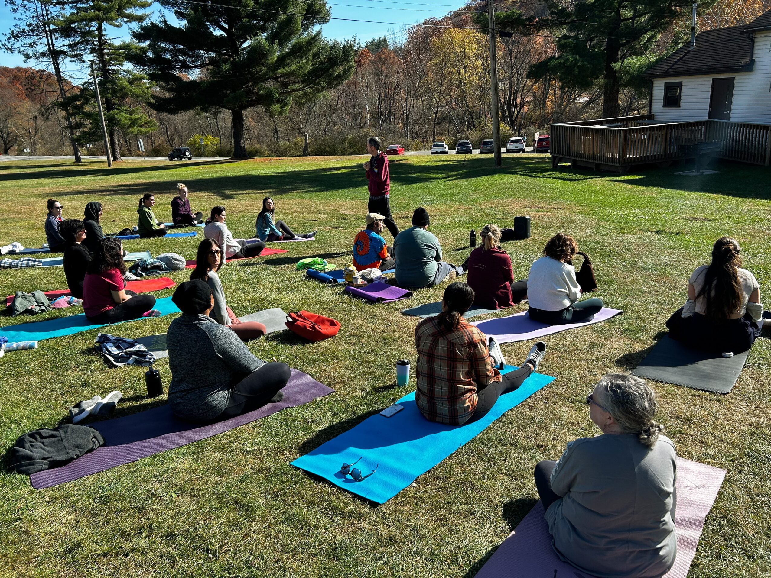 Group of people practicing yoga outdoors on mats in a grassy area with trees and a house in the background.