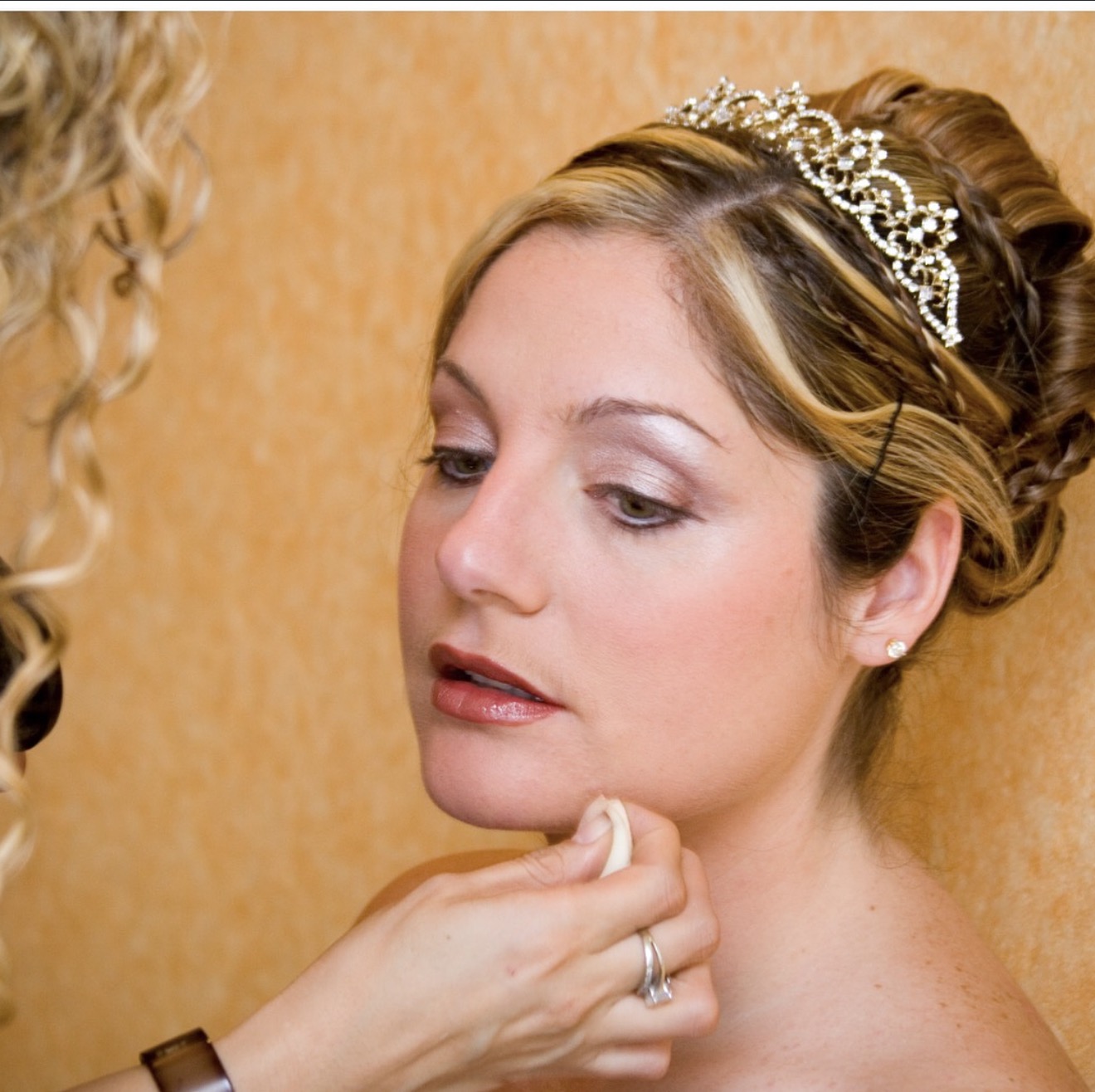 Woman with a tiara having makeup applied, close-up of her face and neck, warm background, detailed hair and makeup, 20 words