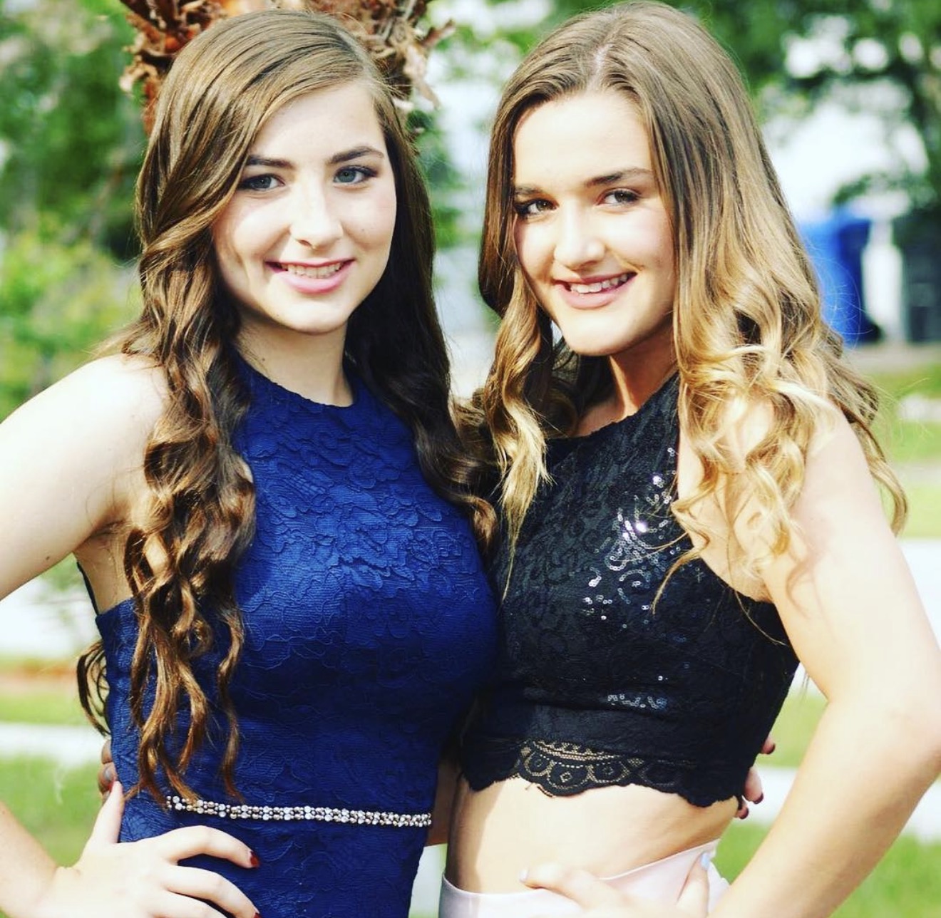 Two young women with long hair smiling outdoors, wearing dark lace tops, standing close together.