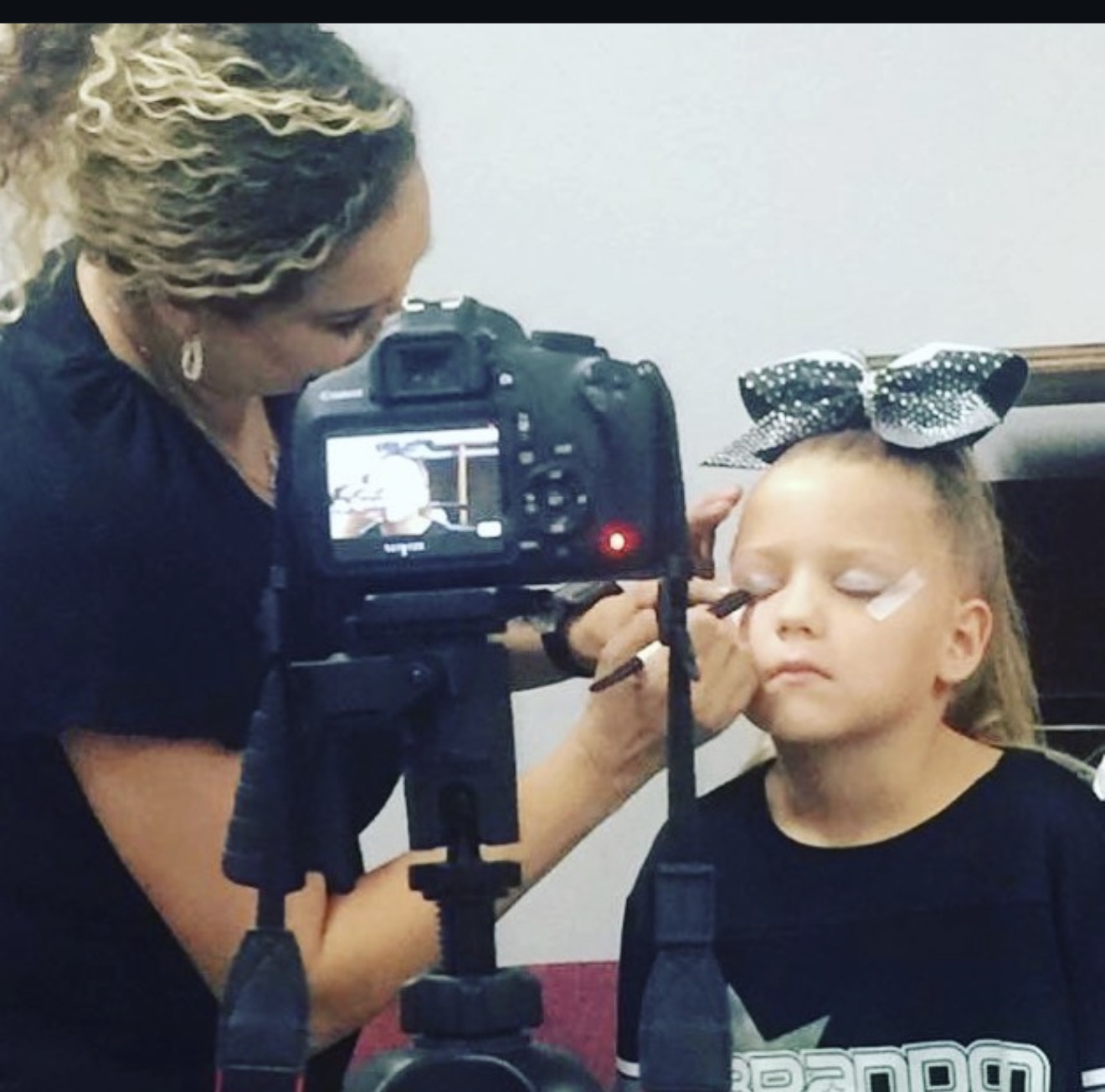 Woman applying makeup to young girl with a camera in foreground, girl has a large bow in hair, neutral background.