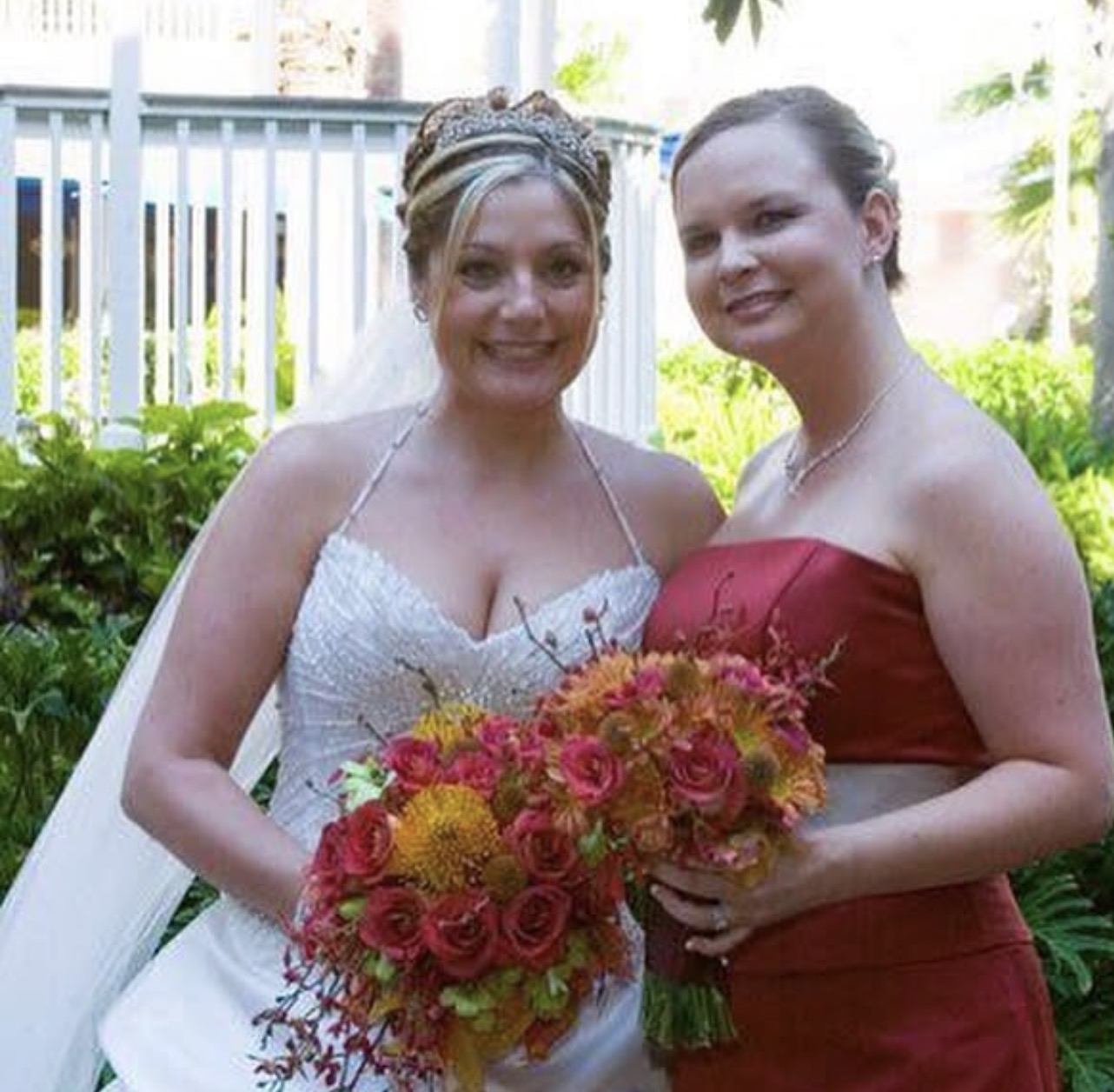 Two women, one in a wedding dress and the other in a red dress, holding bouquets, smiling outdoors with greenery.