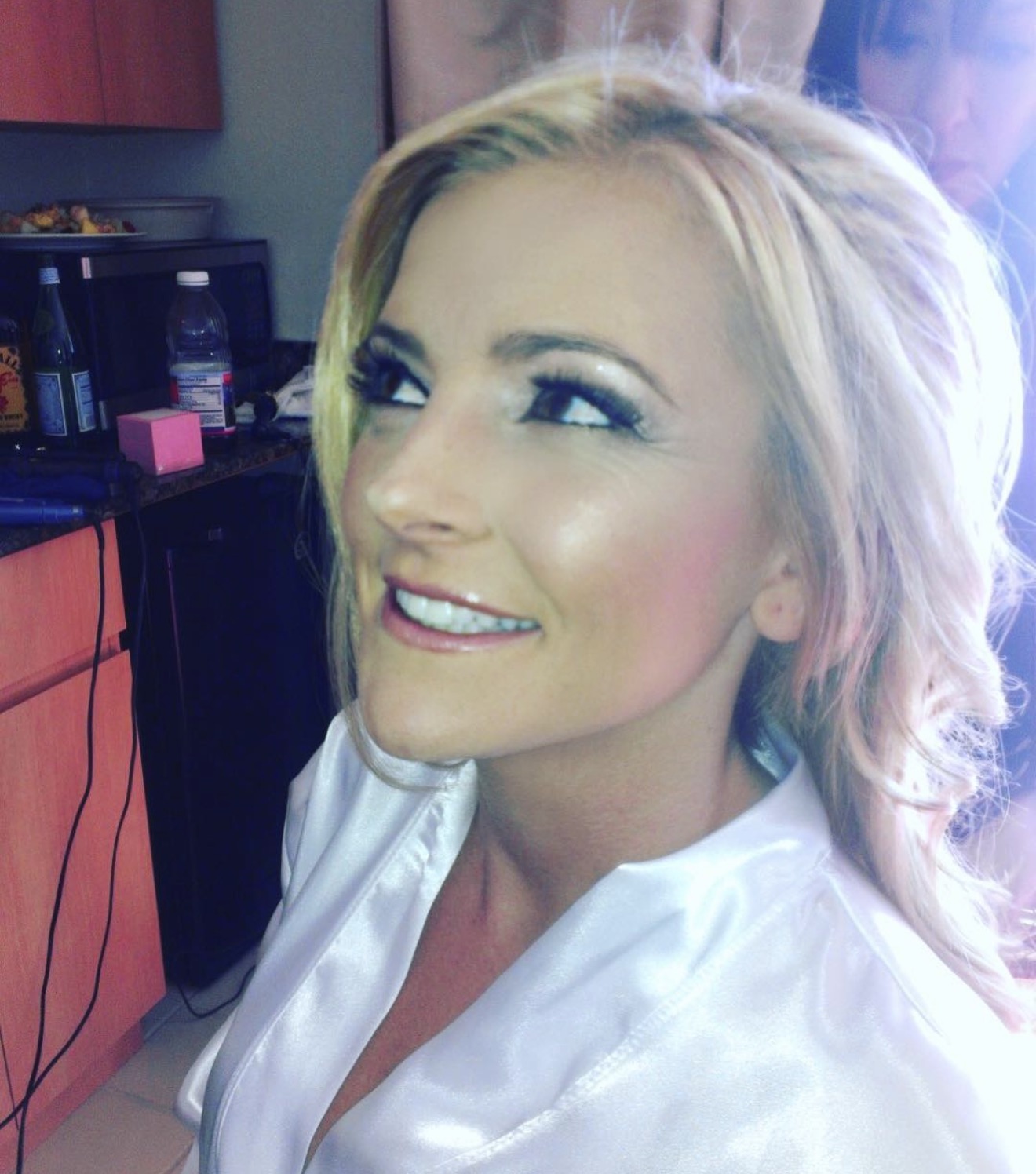 A woman with blonde hair smiling, looking to the side, wearing a white top, in a room with furniture and objects.