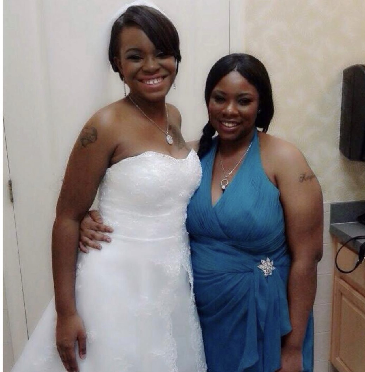 Two women in formal dresses smiling, one in white and one in blue, standing close together indoors.