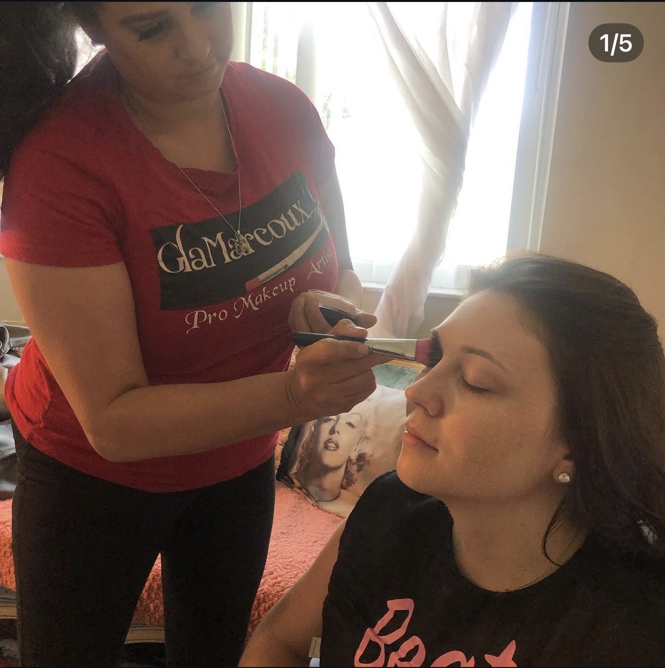 Makeup artist applying makeup to a woman with closed eyes, in a well-lit room with curtains.