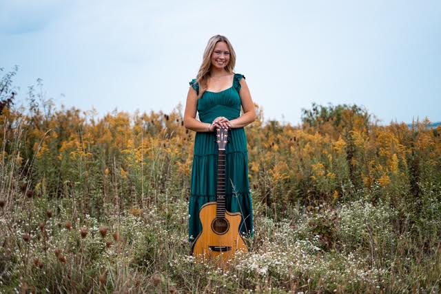 Woman in a teal dress standing in a field with a guitar at her feet, smiling, with yellow flowers and blue sky behind her.