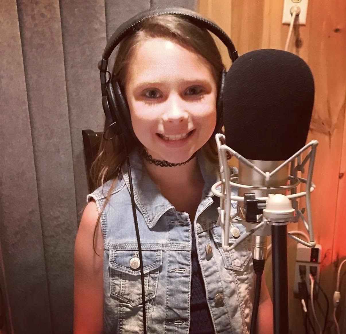 Young girl with headphones and choker in a recording studio, smiling at the microphone.