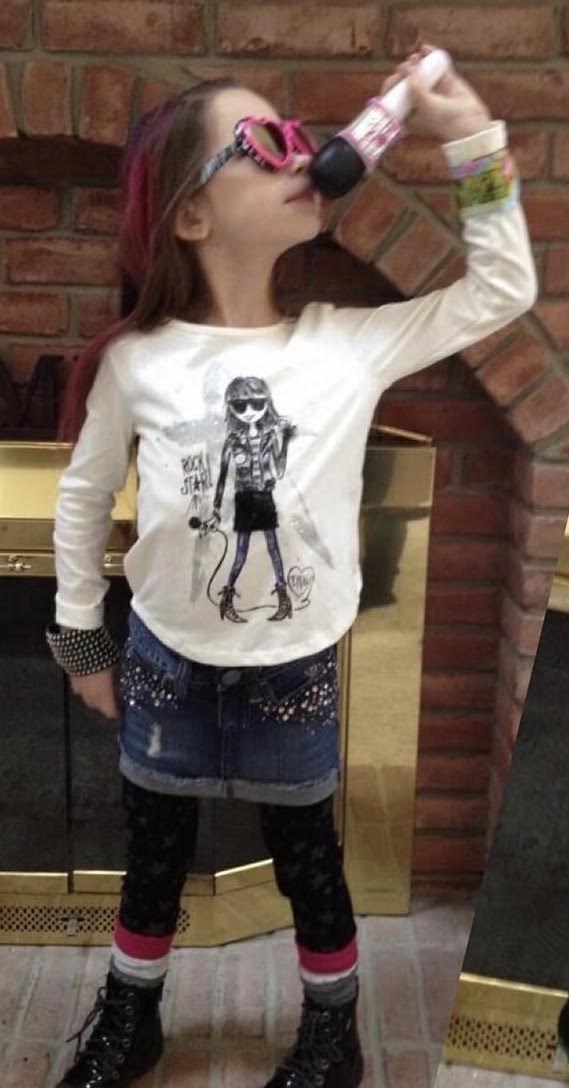 Child drinking from a bottle, wearing glasses, a white sweatshirt, and colorful leggings, standing in front of a brick wall.