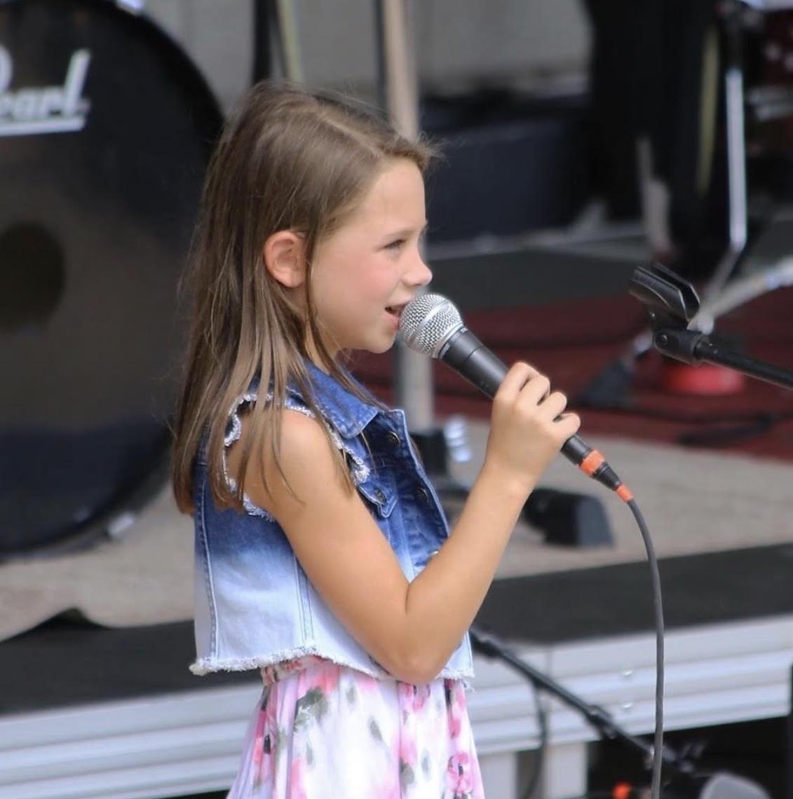 Young girl singing into a microphone on stage outdoors, wearing a sleeveless denim vest and a colorful skirt.